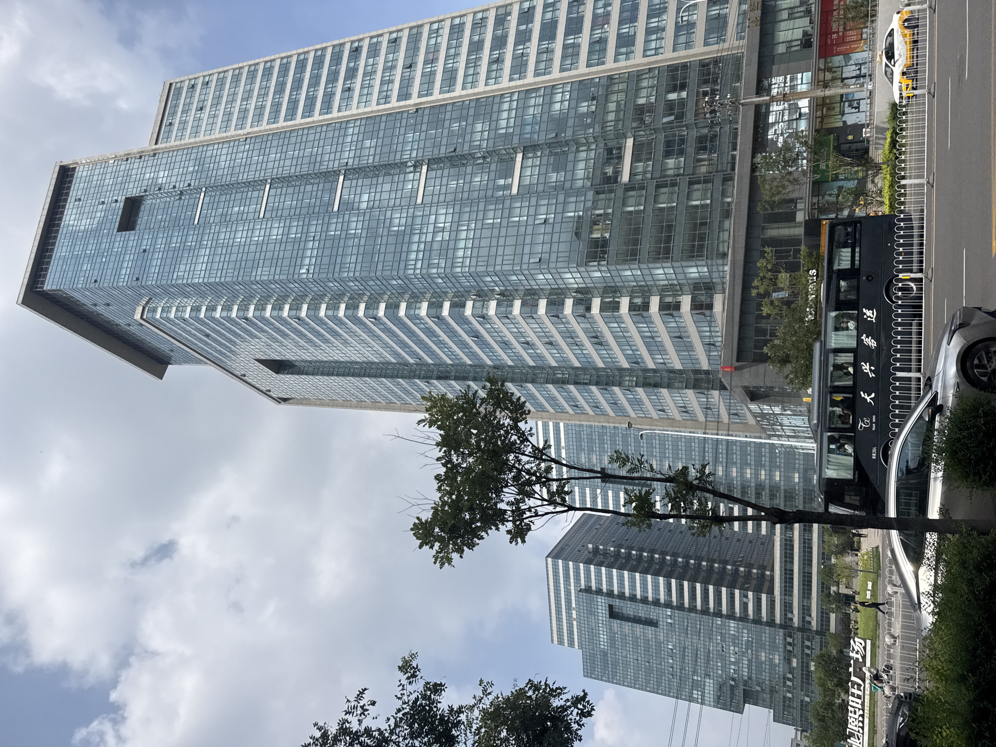 This is a street-level, eye-level, medium shot photograph taken in Dadong District, China. The image captures a modern urban scene dominated by several tall, glass-fronted skyscrapers. In the foreground, a silver car is parked on the right side of the street, partially obscured by green foliage. A black bus with white Chinese characters on its side is stopped in traffic. To the left of the bus, a white sedan with yellow accents, likely a taxi, is also in traffic. Beyond the vehicles, a white metal fence separates the street from a walkway and some greenery. Behind the fence, there is a large sign with Chinese characters, and several people are walking. The background is filled with the imposing architecture of the skyscrapers, with one particularly tall building prominently featured on the right, and two similar buildings to its left, all with many windows. The sky above is partly cloudy, with patches of blue sky visible.
