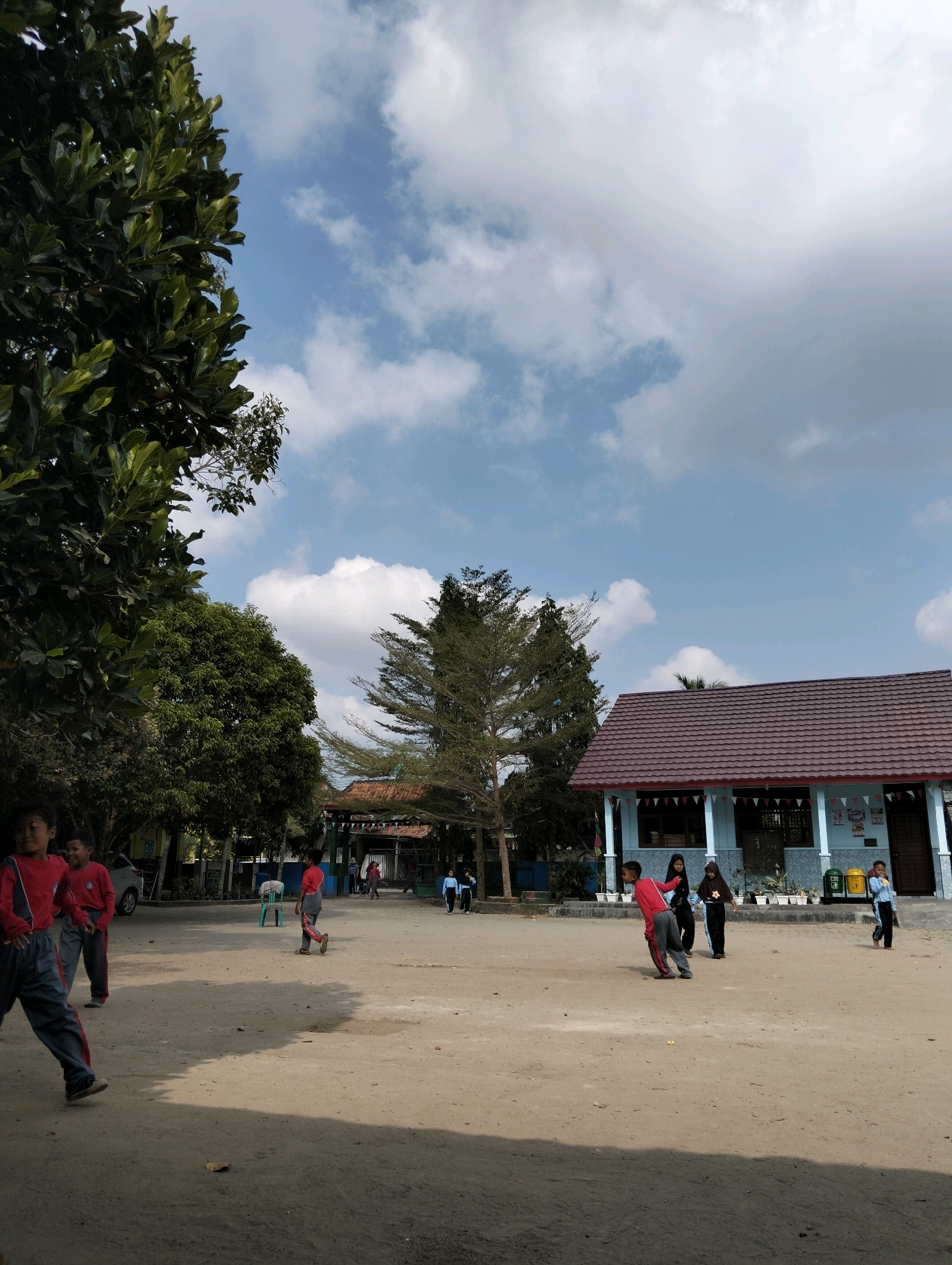 The image depicts an outdoor scene in Banjar Agung, Indonesia, likely a schoolyard, under a partially cloudy sky. In the foreground, a group of children, dressed in casual attire, are engaged in various activities. Two boys in red shirts and gray pants are on the left side of the frame, with one in motion. To the right, another boy in a red shirt and gray pants is walking away from the camera. Further back, a group of individuals, including a person in a red shirt and gray pants and others in darker clothing and headscarves, are interacting. On the far right, a boy in a blue shirt and dark pants is standing. A light blue building with a brown tiled roof, decorated with triangular flags, is situated in the background. Adjacent to the building are trash receptacles and potted plants. Trees are visible on the left and center of the image, providing shade. A silver car is parked on the left. The ground is covered in dirt and sand, with shadows indicating the presence of sunlight.