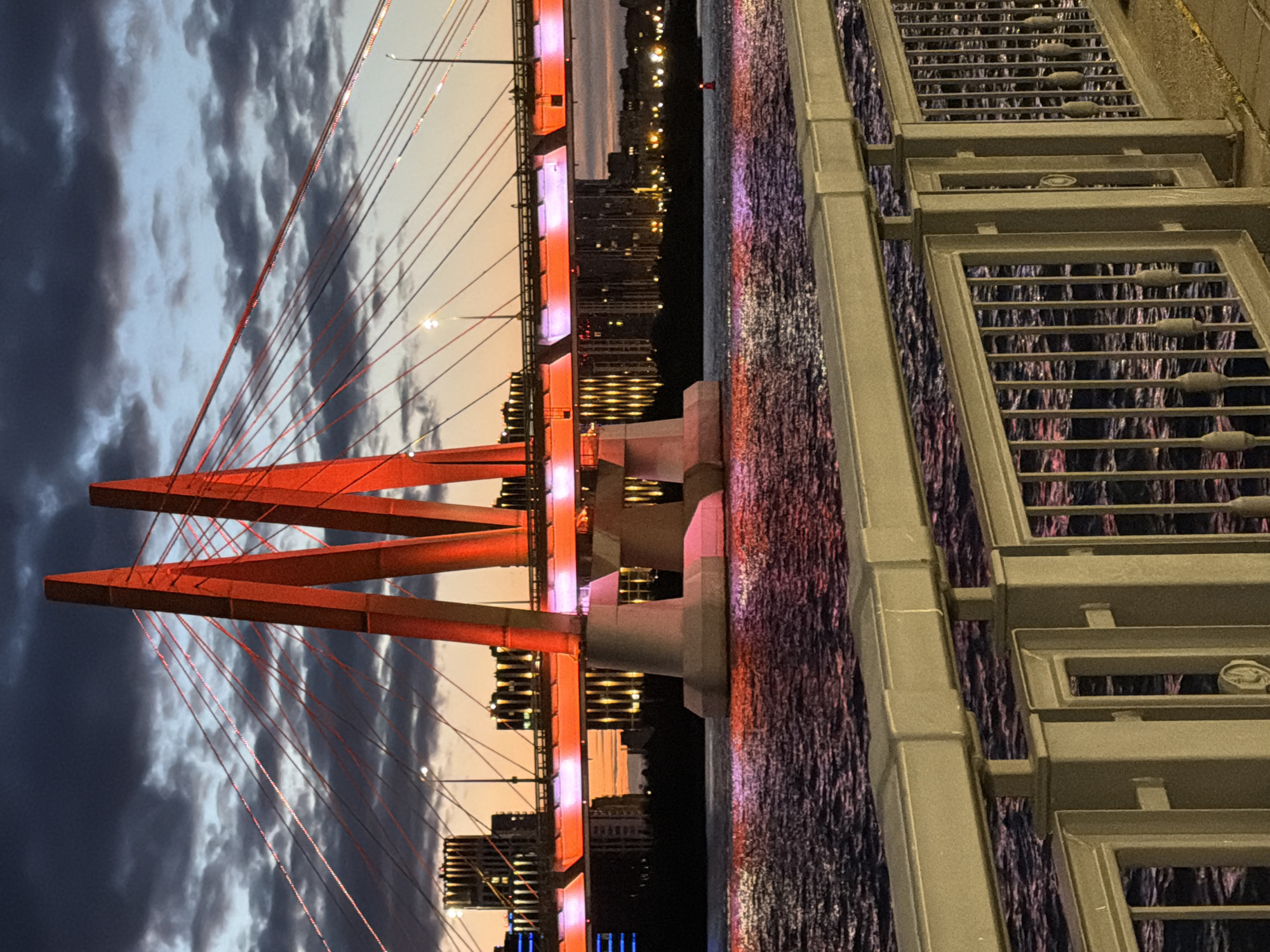 An eye-level, vertical shot captures a modern cable-stayed bridge illuminated with red and orange lights over a body of water at dusk. The sky above is a mix of dark clouds and a pale orange sunset, with a few streetlights visible. The bridge's two massive, V-shaped pylons are lit in bright red, supporting a network of red-lit cables that extend across the frame. The bridge deck itself glows with orange and purple light. In the background, a city skyline is visible with silhouetted buildings and illuminated windows. The water reflects the colorful lights of the bridge and city, creating a shimmering effect. In the foreground, a metal railing separates the viewer from the scene, with a paved walkway behind it. No people are present.