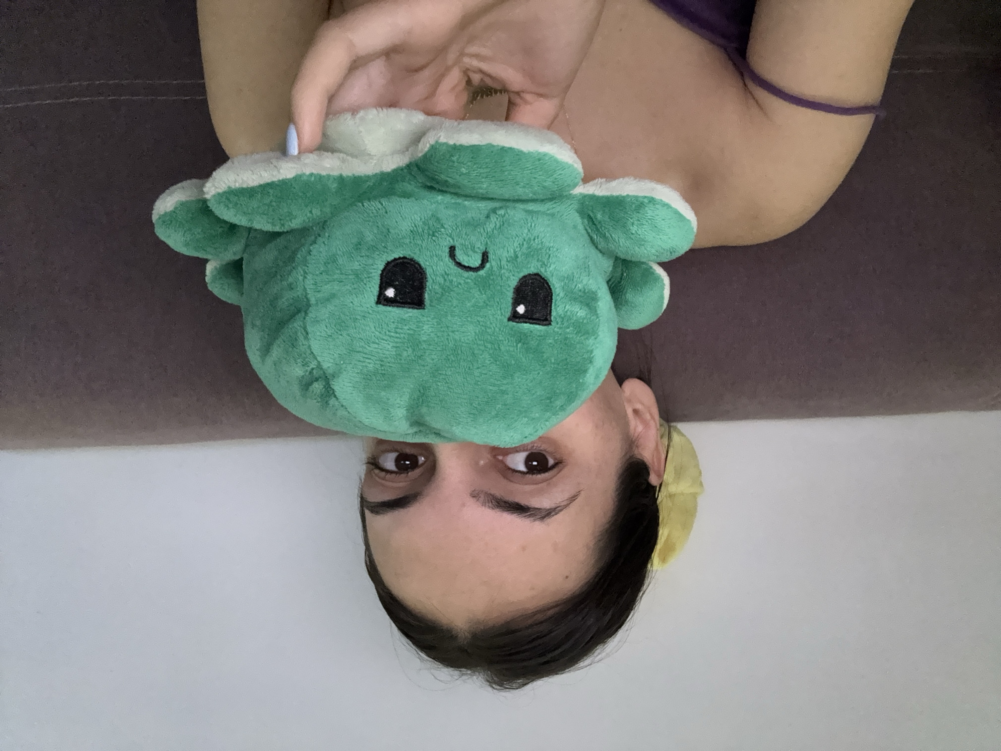 A woman is lying on a brown sofa, holding a green stuffed octopus toy. The woman has brown eyes and dark hair and is facing the camera. She is wearing a yellow hair accessory. The background is a white wall. The location is unspecified and not necessarily in Plovdiv, Bulgaria.