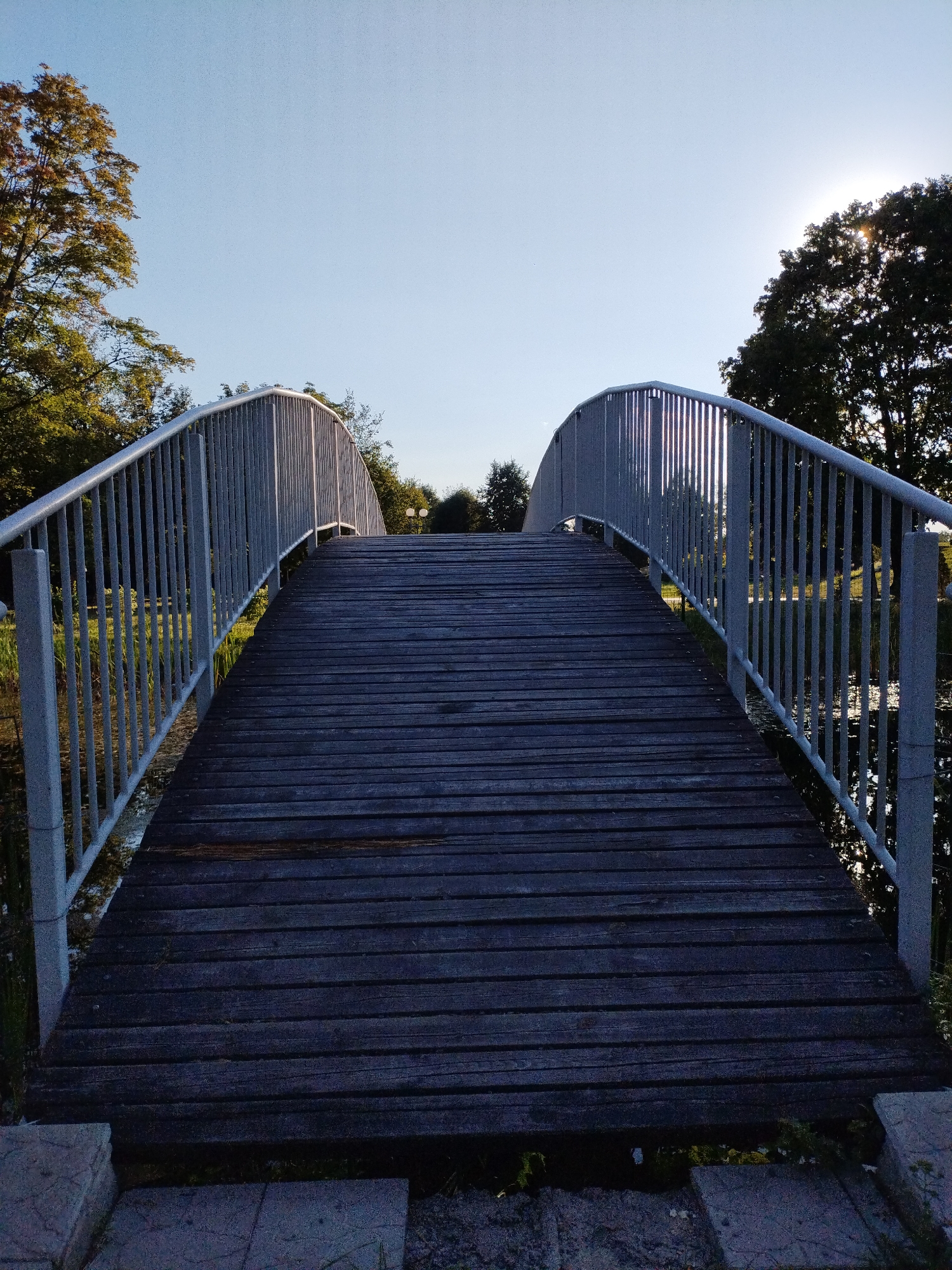 The image depicts a wooden bridge with metal railings arching over a body of water. The bridge is viewed from a low angle, emphasizing its wooden planks and the structure of the railings. Trees with green and autumnal foliage are visible on either side of the bridge, with the sun setting behind the trees on the right, casting a warm glow and creating lens flare. The sky is clear and light blue, suggesting it is late afternoon or early evening. The environment appears to be a park or natural area within the city of Bialynichy, Belarus.