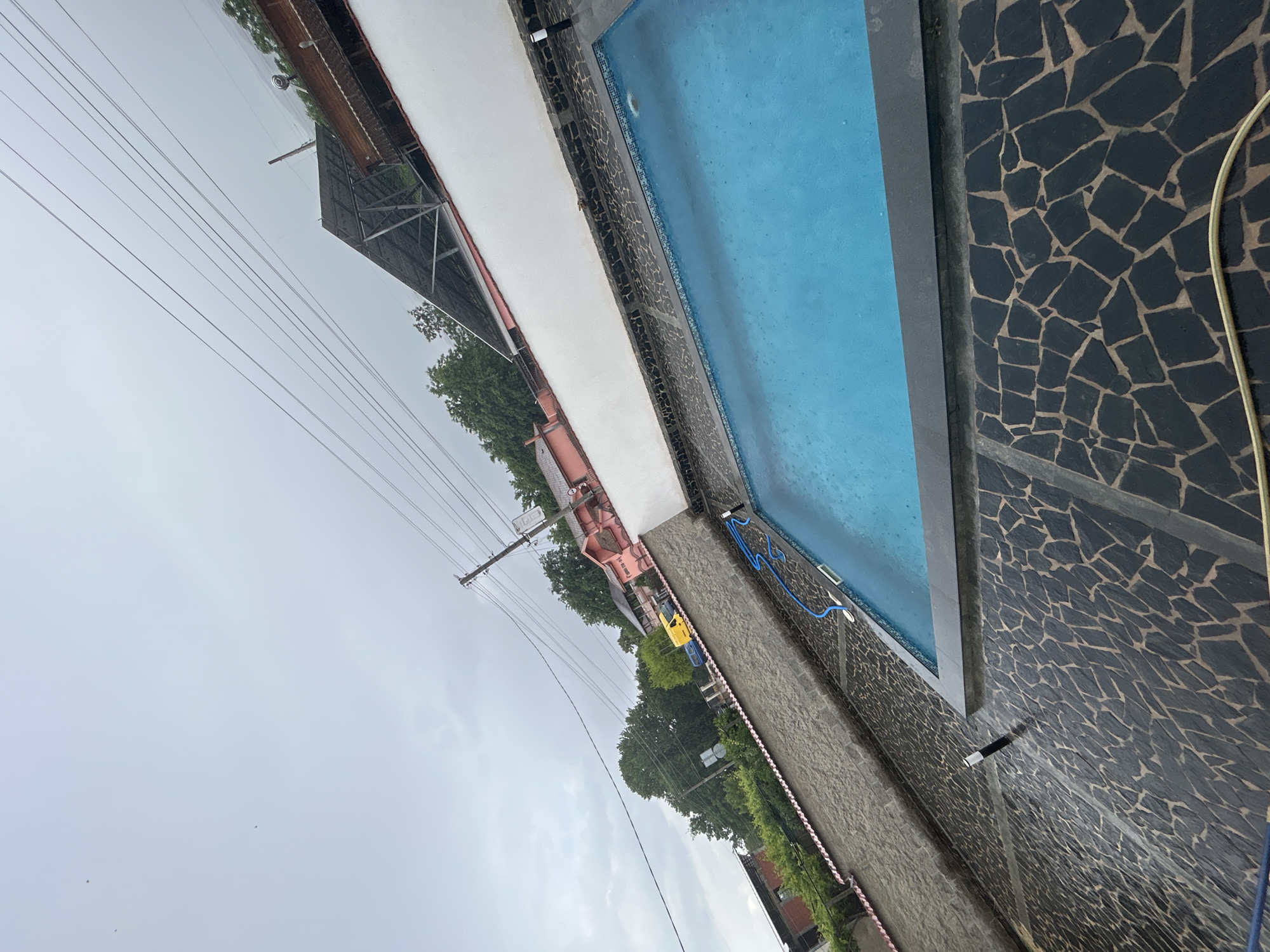 The image, taken in Dolna Gradeshnitsa, Bulgaria, depicts an outdoor swimming pool area under an overcast sky. The main subject is a rectangular swimming pool filled with light blue water, its surface showing ripples, possibly due to light rain or wind.
Alongside one long edge of the pool runs a white concrete-like walkway, which borders a rough, textured grey wall. On the opposite side, the pool is flanked by a dark wall adorned with a distinctive fragmented mosaic tile pattern, consisting of irregular dark grey or black pieces set in lighter grout. A coiled blue hose is placed near the pool on the white walkway side, while a beige hose or cable is visible on the mosaic-tiled side. A black and white cylindrical object protrudes from the mosaic wall.
In the background, beyond the grey wall, are several structures including a building with a red-orange roof and white walls, and a large, dark, open-sided structure with a metallic frame, possibly a canopy or shed. Green trees and foliage are visible throughout the background. Utility poles supporting numerous power lines stretch across the upper portion of the frame, indicating a semi-rural or residential setting. The sky is uniformly grey and cloudy, suggesting a dull or rainy daytime. No people are visible, and no specific activity is clearly taking place. No discernible text is present in the image.