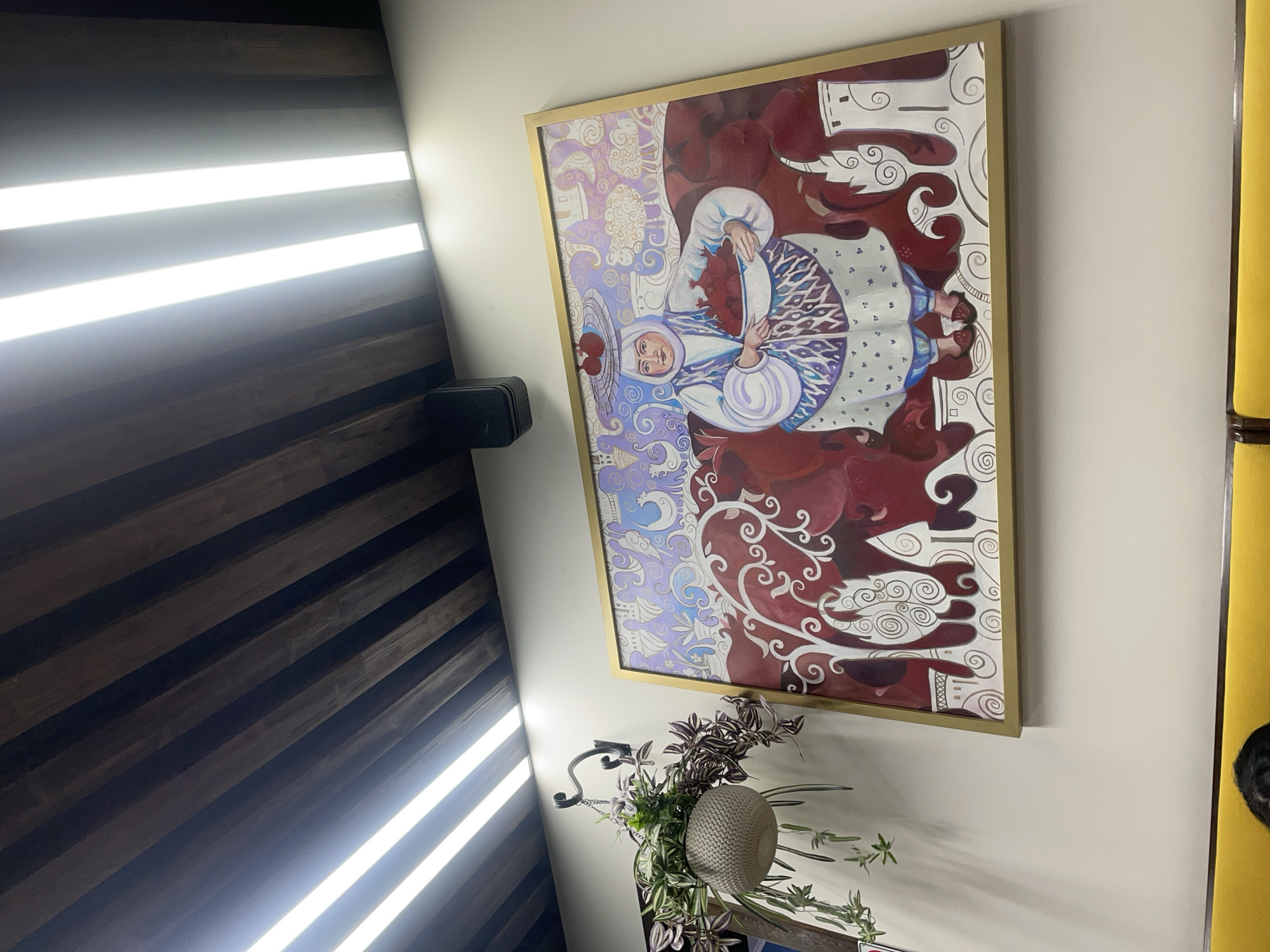 The image shows an interior scene with a large framed painting on a wall. The painting depicts a woman in traditional clothing, wearing a white headscarf and a patterned blue and white dress, holding a bowl of red fruits. The background of the painting features swirling patterns and stylized animals, with prominent red and purple hues. Above the painting, a ceiling is visible with wooden beams and modern LED strip lights. To the left of the painting, a hanging planter with greenery is suspended from a decorative metal hook. The wall is painted a neutral color. At the bottom of the image, a bright yellow upholstered bench is visible, with the back of someone's head and hair showing as they sit on it. The overall setting appears to be a restaurant or cafe in Tashkent, Uzbekistan, given the style of the painting and the presence of traditional motifs.