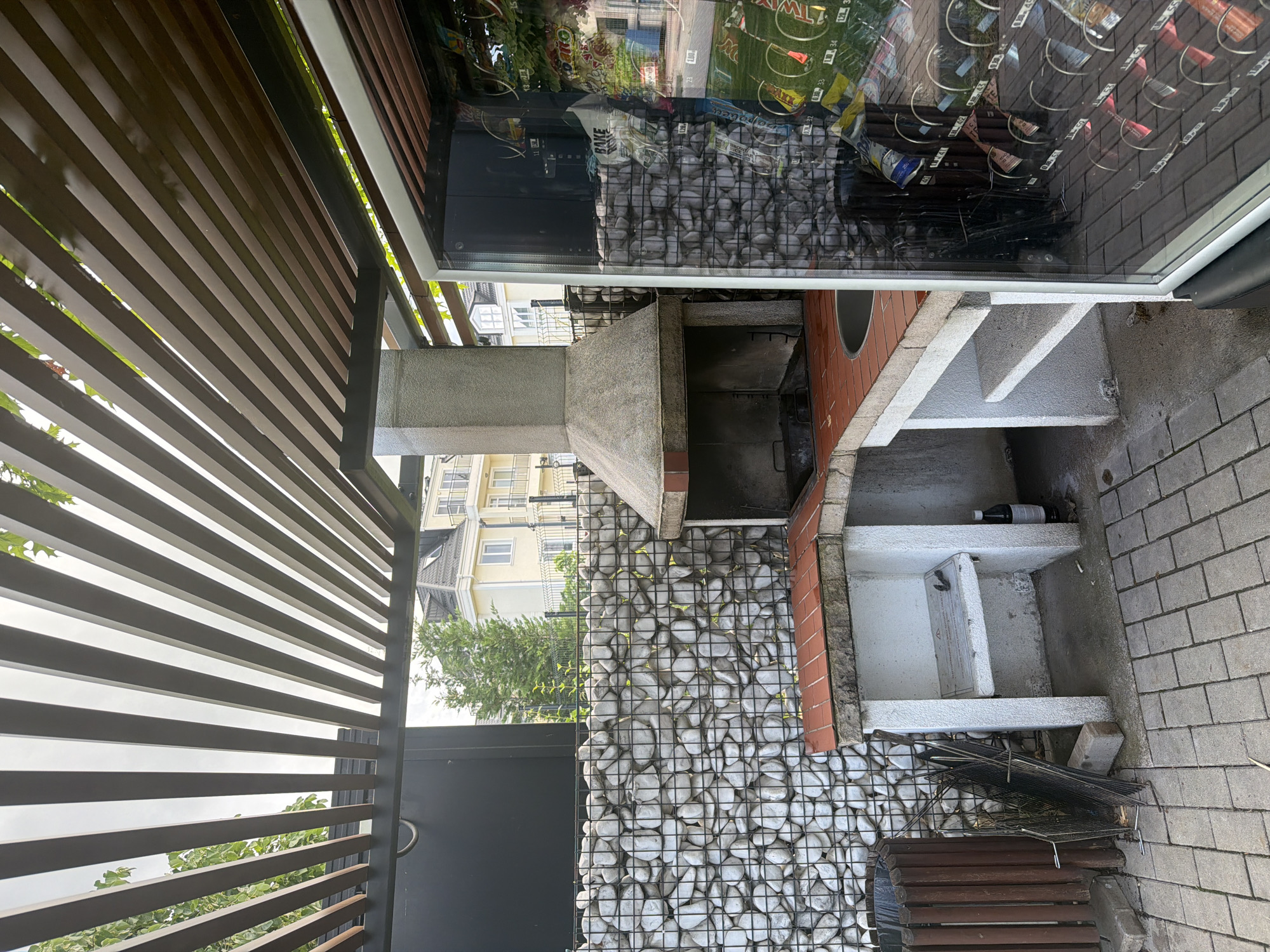 This image captures an outdoor patio or garden area in Sofia, Bulgaria, on a clear, bright day. The scene features a well-equipped outdoor kitchen and leisure space.
In the center of the frame is a robust, built-in BBQ grill with a concrete chimney and a wide grilling area. Adjacent to the grill on its right is a section of reddish brickwork, leading to a concrete countertop with several open shelves underneath. A white bottle rests on the top shelf, and a white basin or sink is visible on a lower shelf. The base of this entire outdoor kitchen unit, spanning the lower-middle portion of the image, is a gabion wall, constructed from wire mesh filled with numerous light-colored stones or pebbles.
The ground area in front of the grill and extending to the right is paved with grey rectangular interlocking blocks. On the pavement in the lower right, a stack of metal grill grates and other dark objects are placed near a small wooden bench or planter.
Overhead, partially obscuring the top-left and upper parts of the image, is a wooden slatted structure, likely a pergola or awning, suggesting a shaded area. Through the gaps in this structure and above the gabion wall in the background, multi-story residential buildings with balconies and windows are visible, interspersed with green trees under a clear blue sky.
A large window dominates the right side of the image, reflecting the surroundings. Clearly visible in this reflection is a vending machine fully stocked with various snacks and confectionery. Recognizable brands include "Twix," "Oreo," "KitKat," "Pringles," "Toblerone," "Ritter Sport," "Milka," and "Kinder." The vending machine also displays item selection numbers such as "11," "21," "23," "31," "41," "42," "43," "51," "52," and "53."
No people are present in the image, indicating the area is currently unoccupied. The scene suggests a functional and well-maintained outdoor space designed for cooking and entertaining.