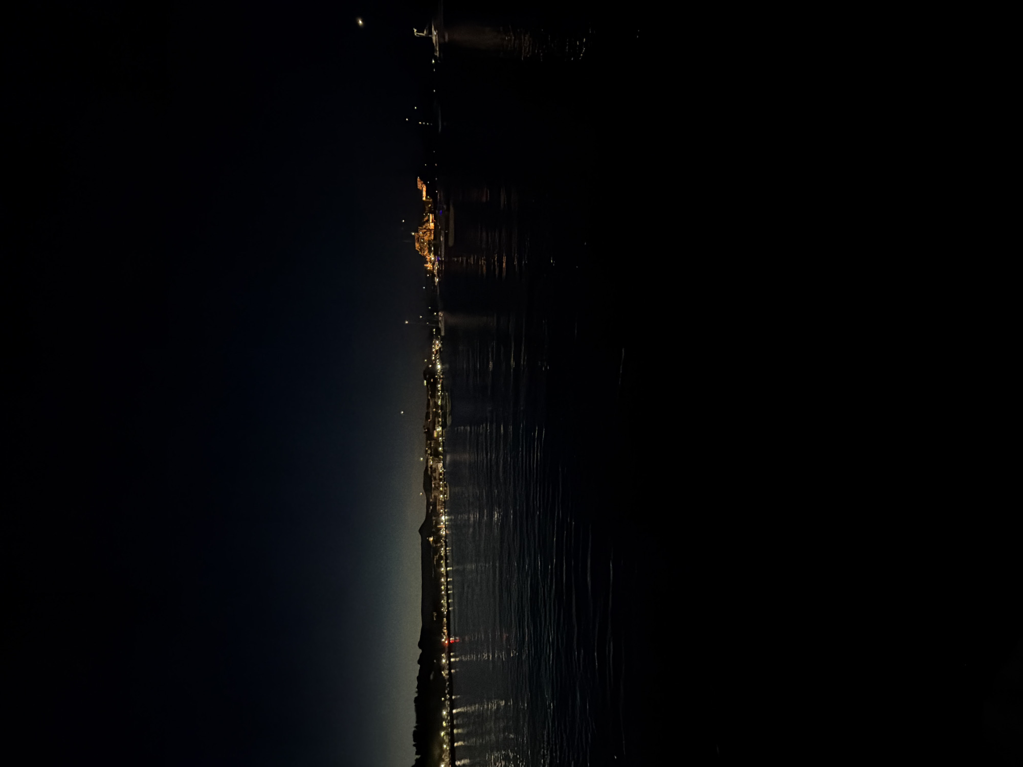 The image depicts a nighttime long-exposure scene over a body of water in Corfu, Greece, specifically facing Corfu Town from the water. The foreground is dominated by dark, undulating water, reflecting numerous artificial lights from the distant shoreline as elongated streaks. Several dark silhouettes of moored or anchored boats, appearing to be yachts or sailboats, are visible on the water, some with faint internal lights.
The midground features the illuminated coastline of Corfu Town. A continuous line of small, bright, yellow-white lights marks the edge of the land, likely from buildings and streetlights along the waterfront. Towards the right-center, the prominent Old Fortress of Corfu is distinctly illuminated with a warm, golden-orange glow, highlighting its fortified architecture and standing out against the darker landscape. Hills and buildings extend behind the immediate shoreline.
The background consists of a very dark, clear night sky, transitioning from a slightly lighter, deep blue near the horizon to an inky black overhead. A singular, small, bright white light source, consistent with a celestial body like the moon or a bright planet/star, is visible in the upper right quadrant of the sky. No individuals or specific actions are discernible in the scene. The overall lighting is low-key, emphasizing the artificial illuminations against the dark environment.