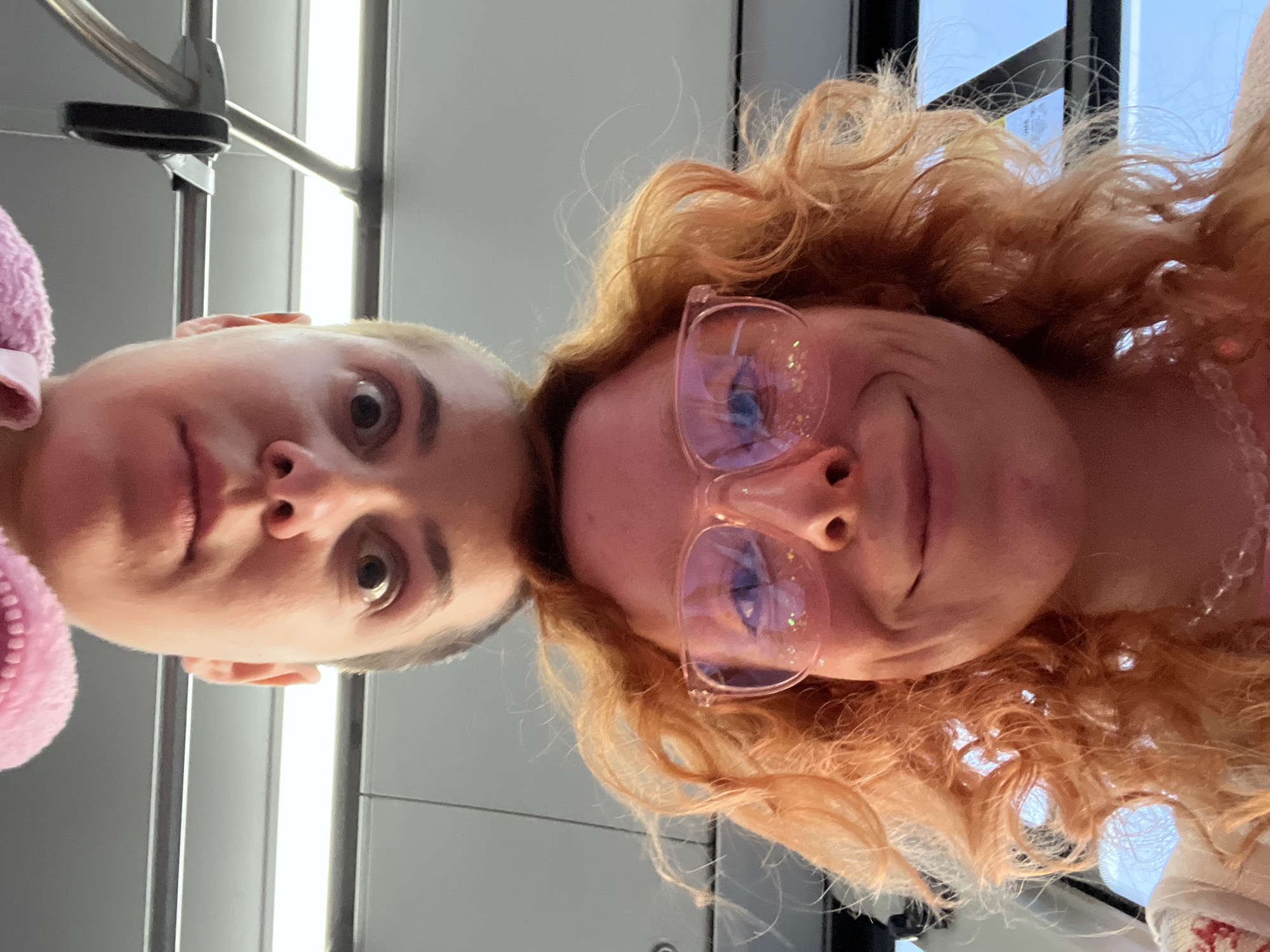 Two individuals are visible in the image. On the left is a person with short, light hair and a pink fuzzy garment visible on the edge of the frame. On the right is a person with curly red hair and pink-rimmed glasses. Both are looking toward the camera. They appear to be indoors. The environment includes light fixtures and metal bars that suggest a train or bus interior. The location in the city of Palilula Urban Municipality, Serbia cannot be ascertained based on the visual information provided.
