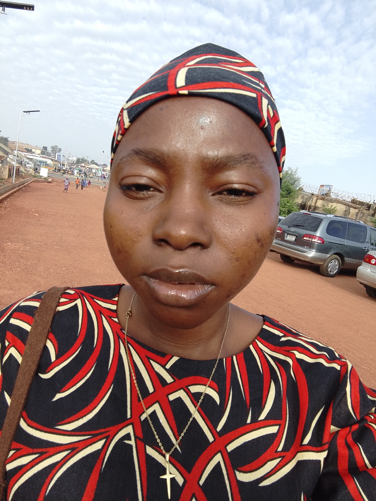 A close-up selfie depicts a woman with dark skin, wearing a patterned head wrap and a matching shirt adorned with swirling red and white designs against a dark background. She is also wearing a gold necklace with a cross pendant.
The backdrop indicates an outdoor setting, likely a city street in Gura Top, Nigeria. A red dirt road is visible, with a few indistinct figures walking in the distance. In the background, a grey minivan and parts of another car are parked near a low wall topped with barbed wire. Utility poles are also visible, along with other buildings and structures indicating an urban or semi-urban environment. The sky is overcast, with scattered clouds.