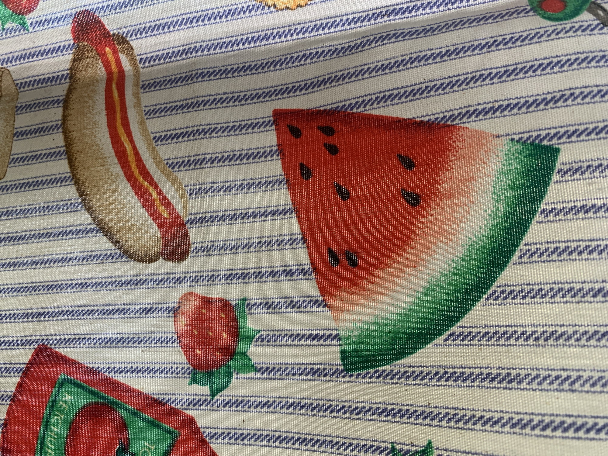 The image shows a patterned fabric with a light background and blue horizontal stripes. Printed on the fabric are depictions of food items, including a hot dog, a slice of watermelon, and a strawberry. There is also a partial image of a ketchup bottle. The fabric appears to be textured and is not lying flat. The scene is a close-up of the fabric itself, without any identifiable location in Santa Ana, Venezuela.
