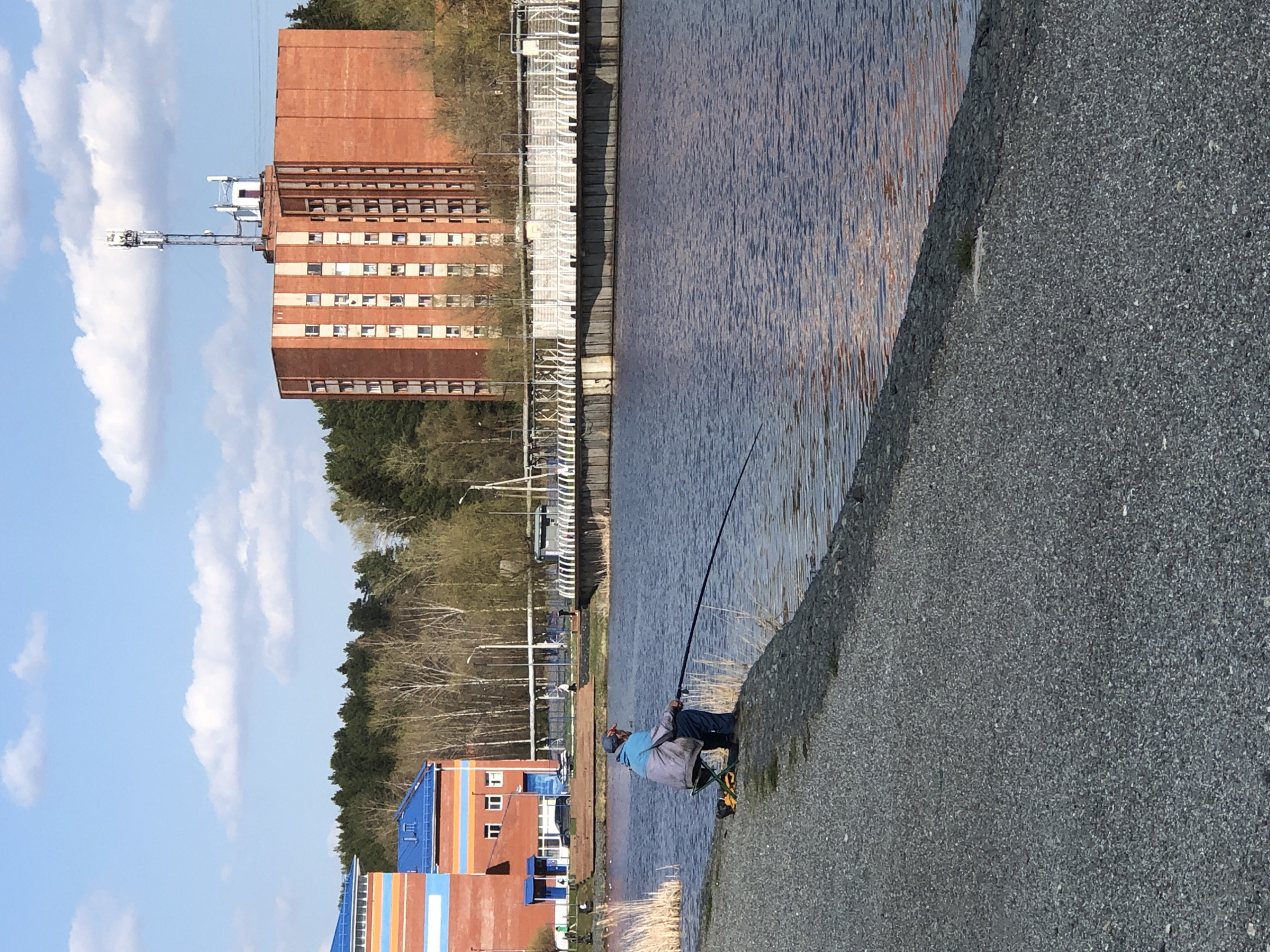 A man sits on a folding chair next to a body of water, holding a fishing rod. The shore is made of gray, small gravel. Across the water, there is a red brick building with numerous windows and a white communication tower extending from its roof. To the left of the building is a line of trees, and further left, another building with blue and red stripes. The sky is pale blue with scattered white clouds. The setting is the city of Votkinsk, Russia.