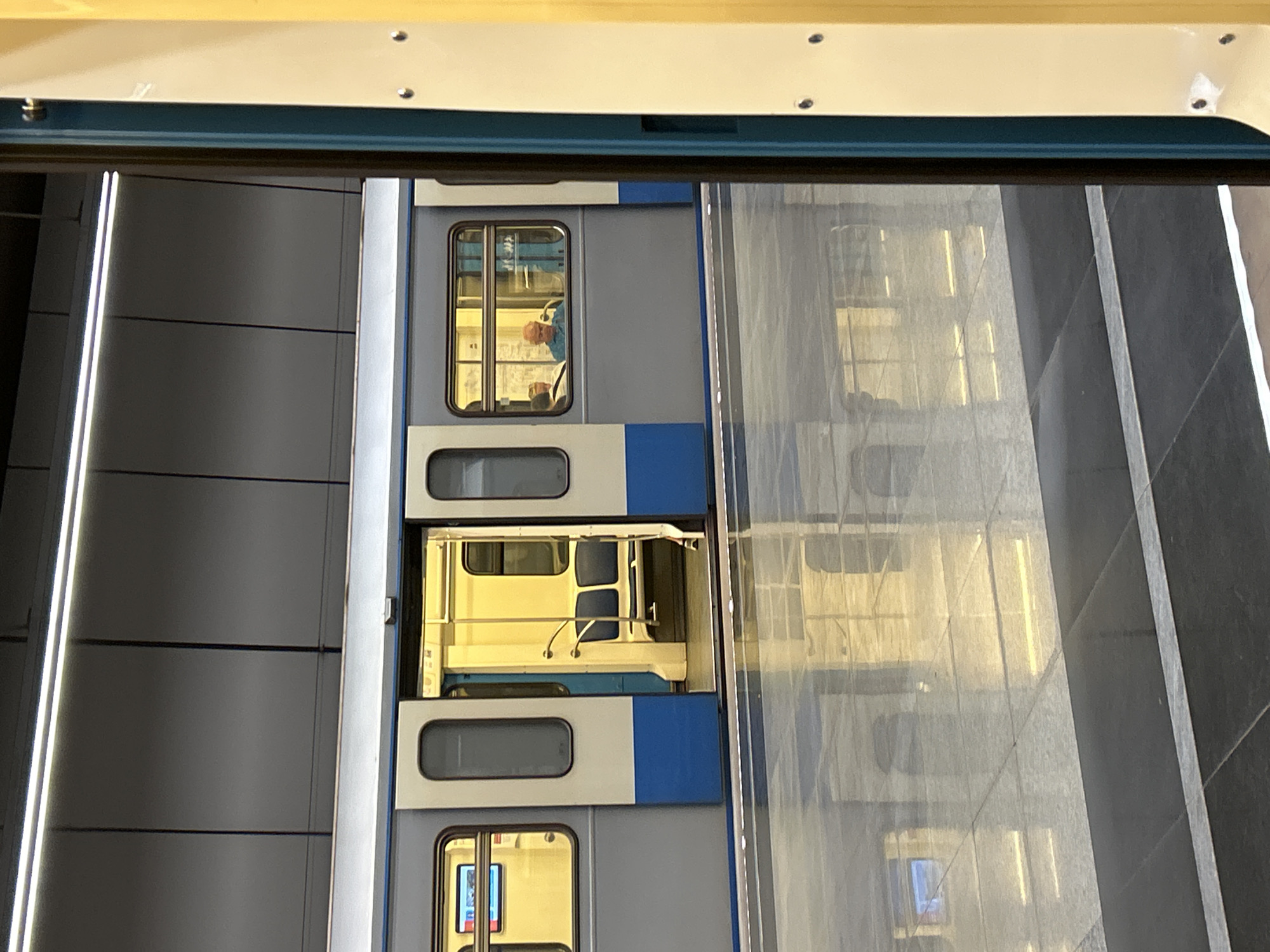 The image shows a subway train stopped at a station platform. The train is gray and blue, with open doors revealing empty seats. A man with a bald head and a blue shirt is sitting by a window on the train. The platform is made of reflective tiles, with the lights from the station ceiling reflecting on the floor. The station appears to be modern and well-lit.