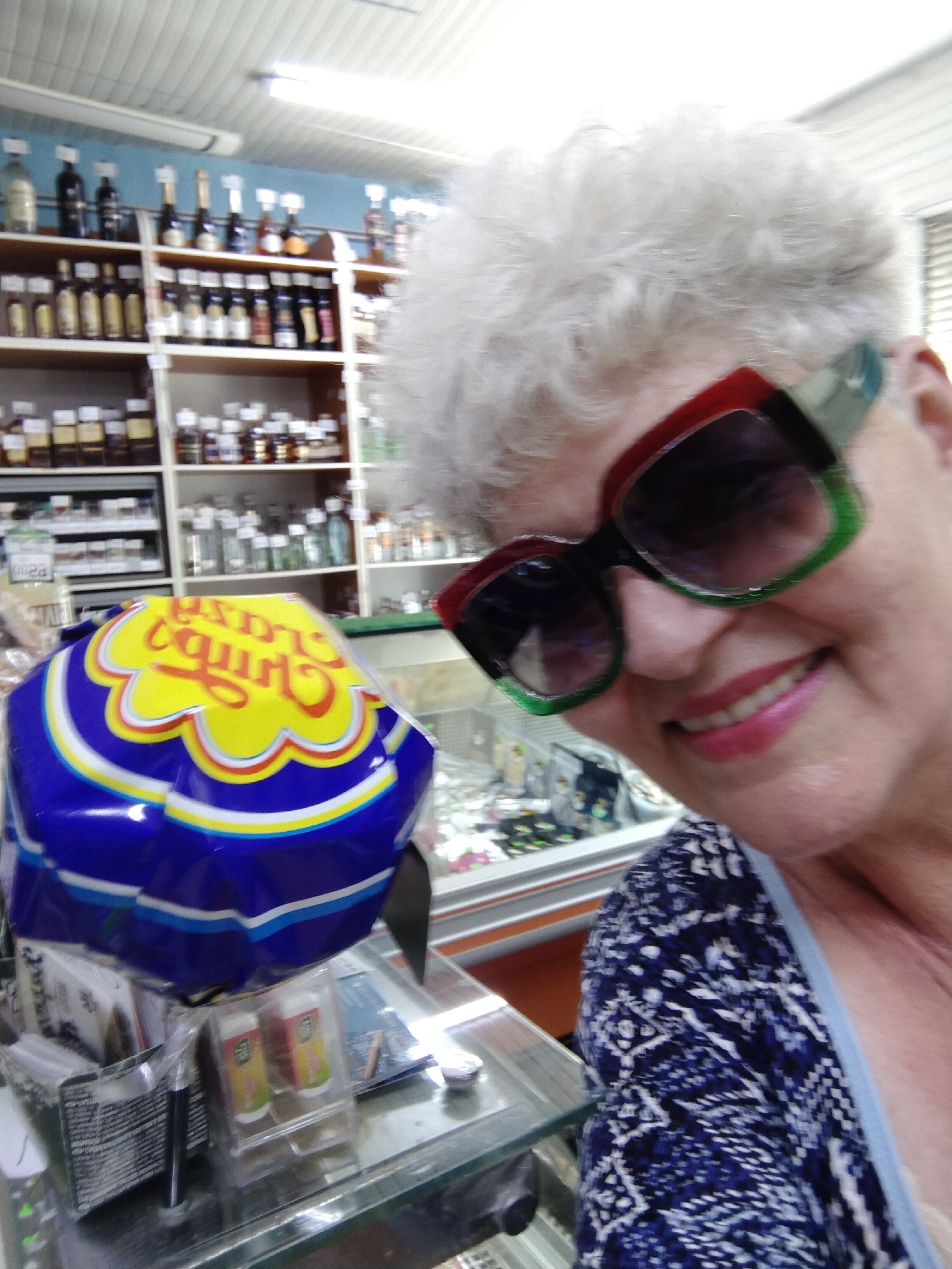 In a retail environment, likely a convenience store or supermarket, a woman with short, white, curly hair, wearing striking oversized sunglasses with red and green frames and dark lenses, is captured in a close-up, selfie-style shot. She is smiling, her lips painted a vibrant pink. She is wearing a dark blue and white patterned top. In the foreground, a large, brightly colored bag, possibly containing snacks, is prominently displayed. The bag features a yellow and red design with blue accents. Behind the woman, shelves stocked with various bottles and products are visible, suggesting a wide array of merchandise. The overall setting appears to be a well-lit indoor space with a tiled ceiling. The store is located in the city of Horishni Plavni, Ukraine.