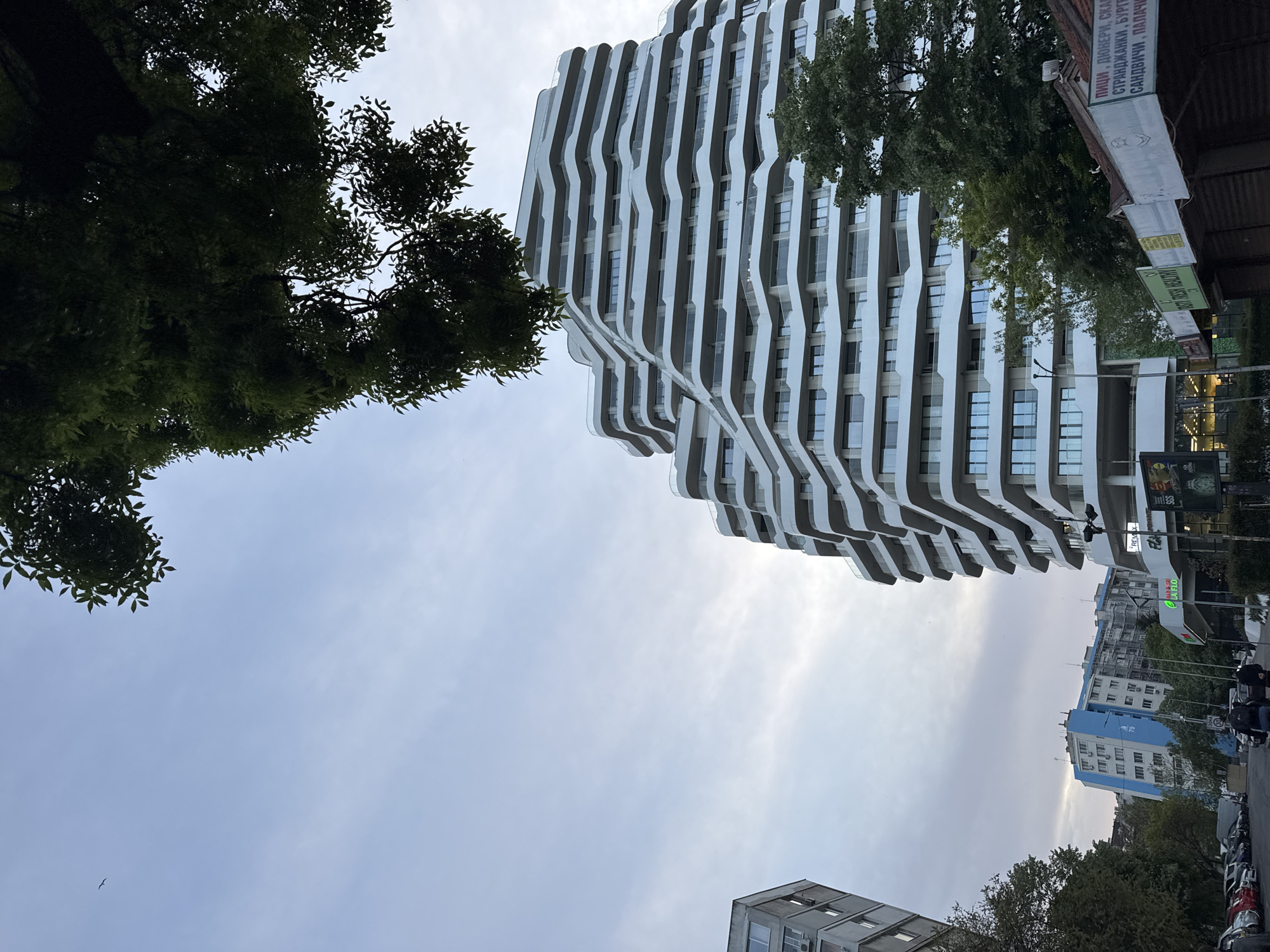 The image depicts a view upward towards a modern, multi-story building with a distinctive wavy, white façade, located in Burgas, Bulgaria. The sky is overcast with light grey clouds. A tree with dense, dark green foliage is visible in the top left corner. The top right of the frame includes a section of a building with signs in Cyrillic. In the distance, a blue and white building can be seen. The image is rotated counterclockwise.