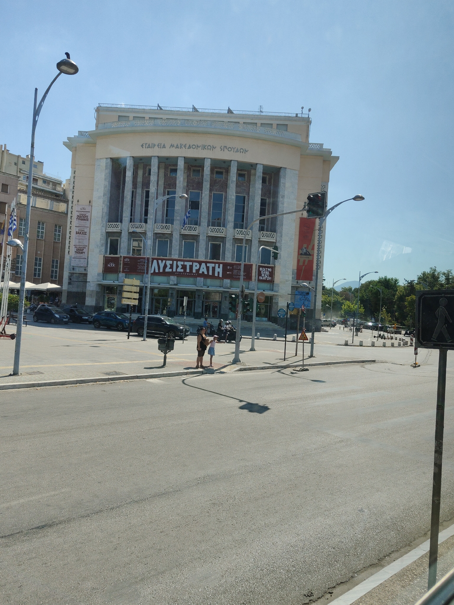 This image shows a street scene in Thessaloniki, Greece, featuring the Thessaloniki State Theatre of Ancient Drama. The building has a classical facade with columns and is adorned with banners and signage. In the foreground, the street is paved and appears to be a multi-lane road with a pedestrian crossing. Several cars are parked or driving on the road. Pedestrians are visible on the sidewalk, including two individuals standing near a traffic light pole. The sky is clear and blue. The environment suggests a sunny day in an urban setting.