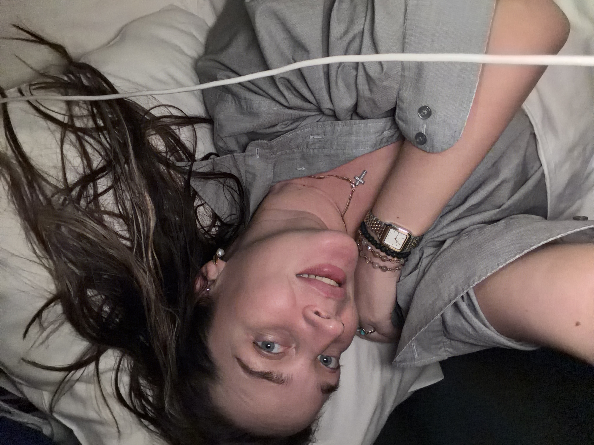 A woman with brown hair and blue eyes lies on a white pillow in a bed. She wears a gray button-down shirt, a watch, bracelets, and a cross necklace. A white wire is visible across the top of the image. There is no indication of the location being Moscow, Russia.