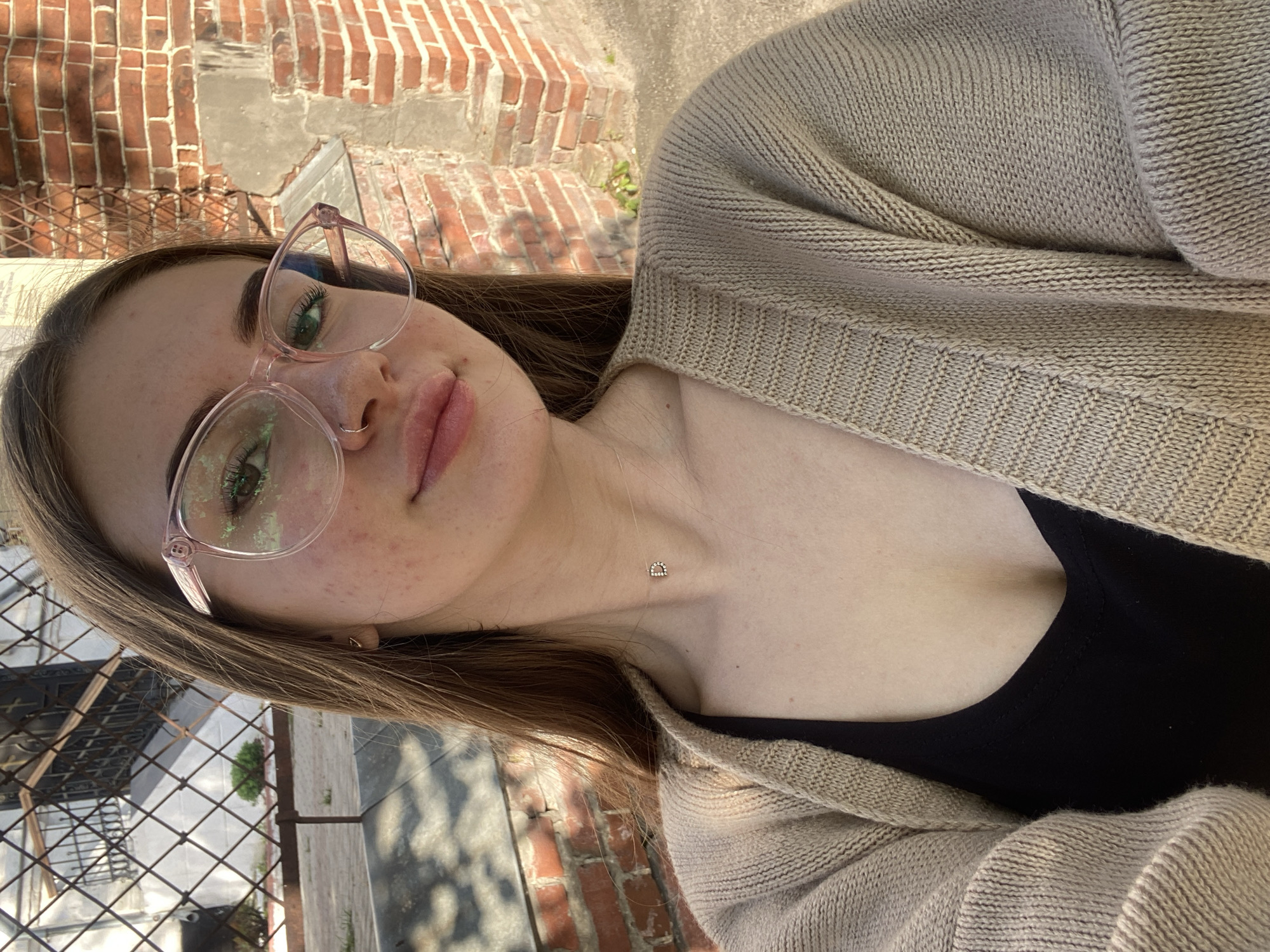 A close-up, upward-angled selfie captures a young woman, displaying her upper body and face. She has light brown hair and green eyes, partially visible through contemporary pink-framed eyeglasses. Her lips are slightly parted, revealing a natural expression, and a small, delicate nose ring adorns her left nostril. Her skin shows some natural texture and minor blemishes on her cheeks.
She is dressed in a light beige or cream-colored knitted cardigan worn over a simple black top. A small, sparkling, gold-colored necklace, shaped like the letter 'd' or a stylized horseshoe, is visible on her collarbone.
The background indicates an outdoor, sunlit urban or semi-urban setting. To her left, a dark metal fence or grating is present, while a textured red brick wall with visible mortar lines occupies the space behind and to her right. The bright light suggests it is daytime, casting clear illumination on the scene. The woman looks directly at the camera, conveying a relaxed and composed demeanor while taking the photograph.