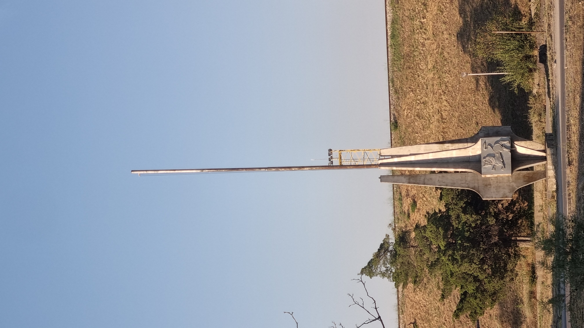 The image shows a tall, slender monument with a metallic top, located in the city of Ruse, Bulgaria. The monument features two abstract concrete pillars forming a base, with a decorative metal plaque depicting a lion on the front. Behind the monument, a sloped grassy embankment is visible, with a train track running along the top. To the left of the monument, a mature tree with dense green foliage stands. The sky is clear and blue. The monument is situated in an open area, likely a public space or park, with sparse vegetation and a paved surface in the foreground. The overall impression is one of an outdoor public monument set against a natural backdrop.