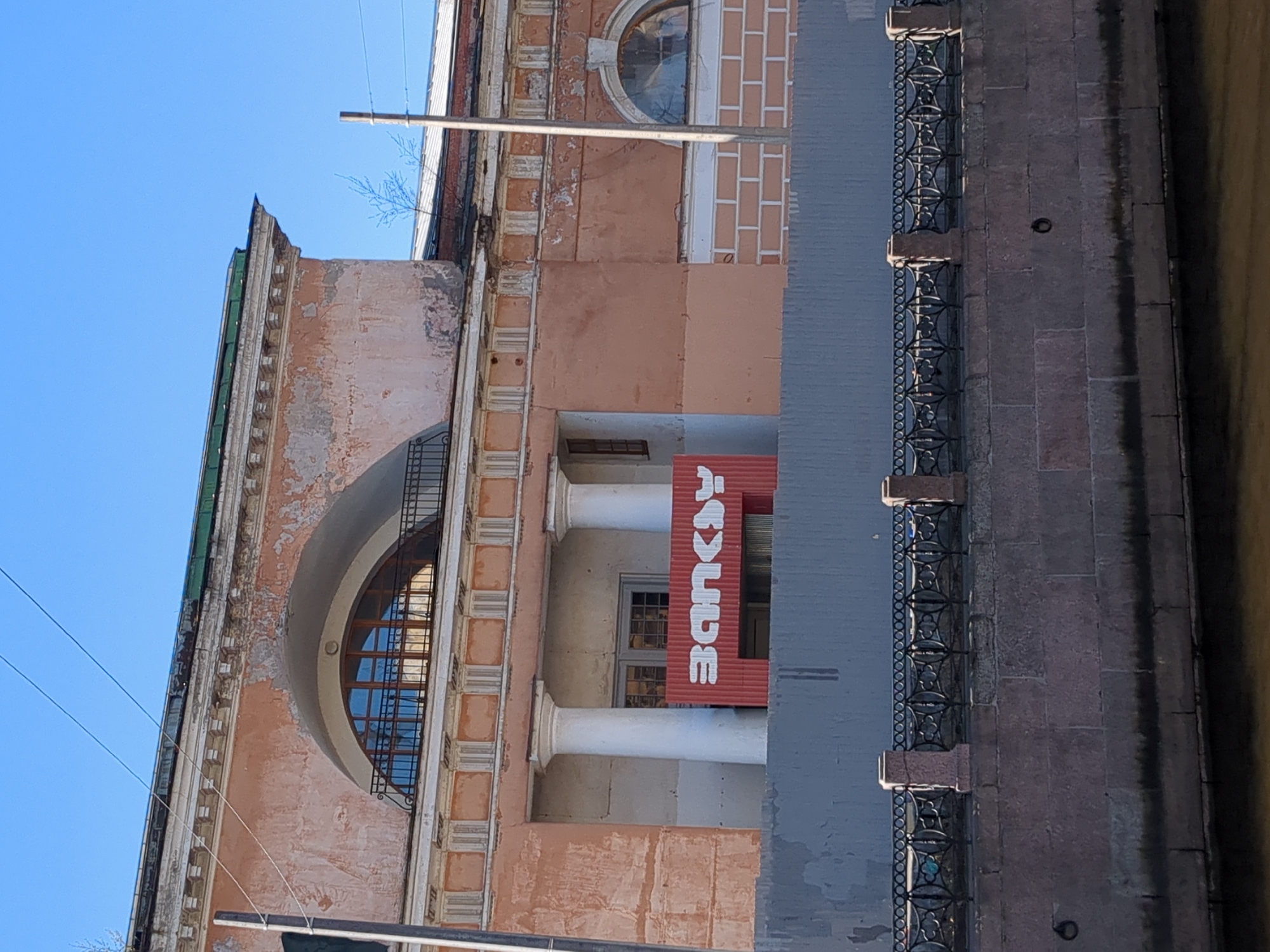 The image shows a building in Saint Petersburg, Russia. The building is old, with a pink facade and decorative architectural details such as columns and arched windows. A red sign with the word "ROUGE" in white letters hangs from the building. A gray wall and a metal railing run along the bottom right of the image, suggesting the building is located next to a canal or river. The sky is blue.