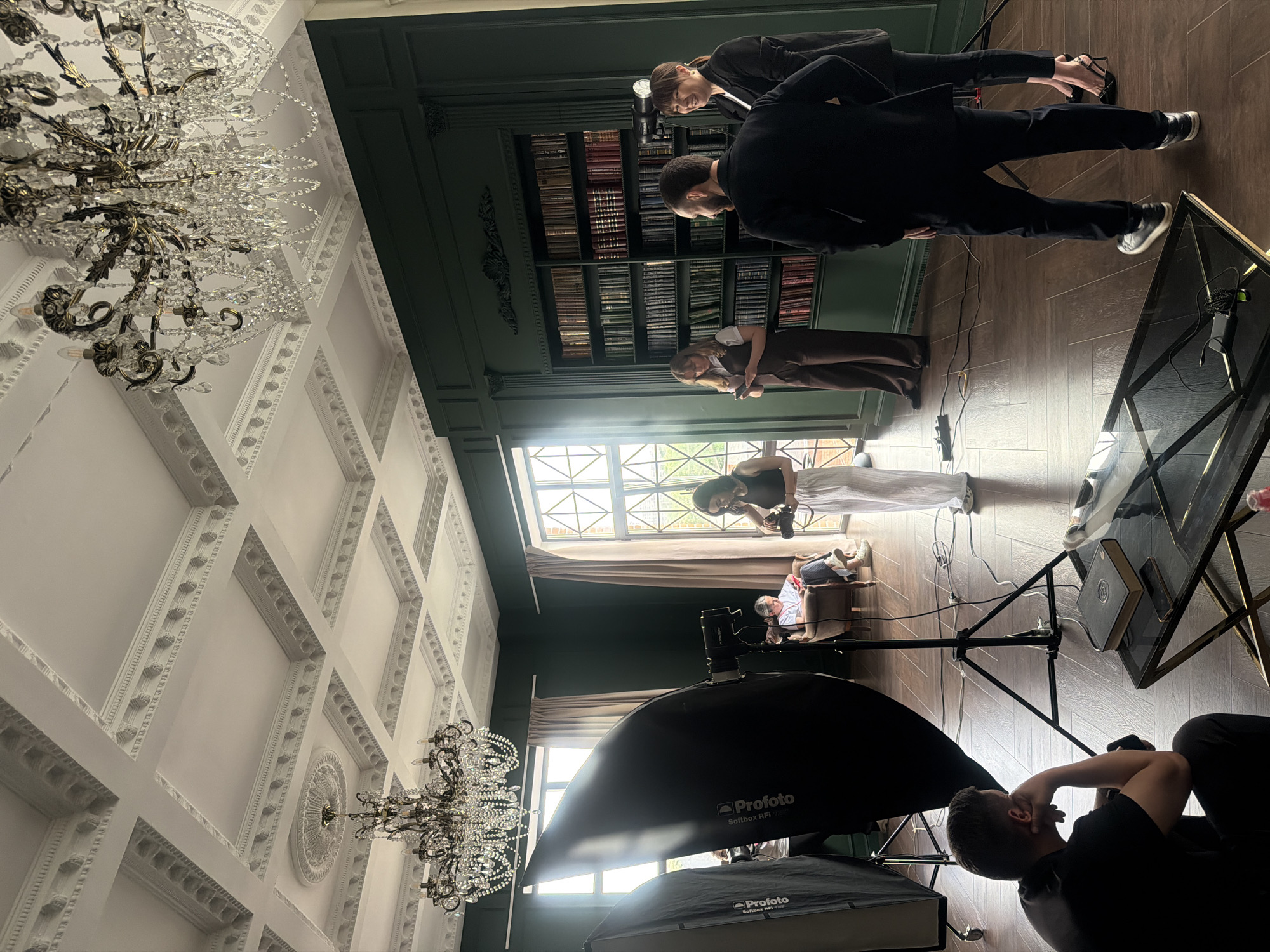 The image depicts a photoshoot in what appears to be a grand room, likely in Rostov-on-Don, Russia. The location suggests this city because Rostov-on-Don is a well known city in Russia. The room has high ceilings with intricate white moldings and large chandeliers. One wall is covered with what appears to be a dark green or black paneled wall and built-in bookshelves filled with books.
The scene involves multiple people: a person sitting and being photographed, a person operating a camera, and other people standing around possibly involved in the shoot. Photography lighting equipment is prominently visible, including large softboxes by "Profoto." Various objects, such as a glass-topped table, wires, and books are scattered around the floor. The focus of the shoot seems to be the person seated in a chair near a large window with light streaming in. The overall impression is that of a professional photoshoot taking place in an upscale interior.