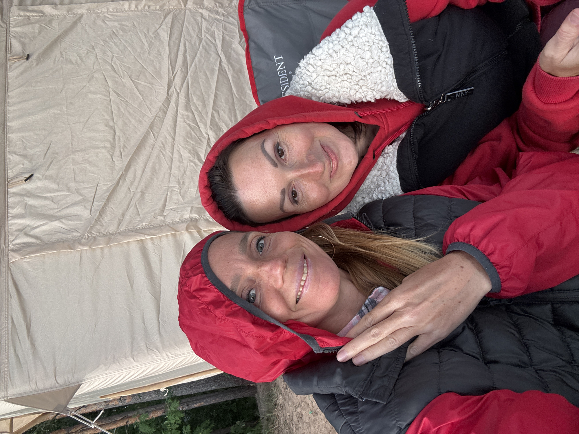 Two women are taking a selfie outside a tent. The woman on the left is wearing a red hooded jacket and a black vest. She has blonde hair and is smiling. The woman on the right is wearing a red hooded sweatshirt with a white sherpa collar. She has brown hair and is also smiling. Both women are wearing red outerwear. The background is a beige tent. The scene appears to be in an outdoor setting, possibly a campsite. There is no visible indication of the specific location in Shepelevo, Russia.