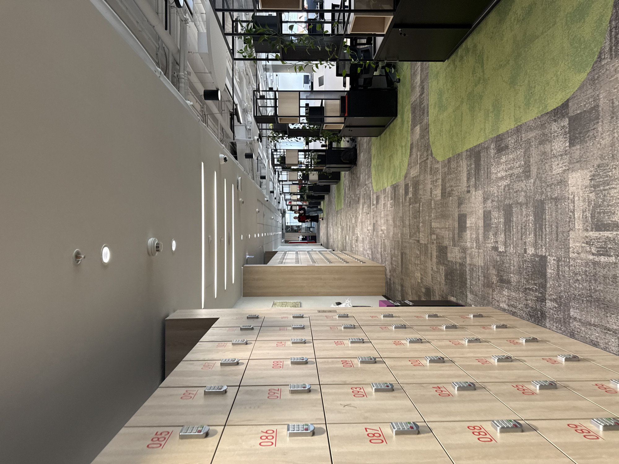 Here is a description of the image:
The image shows a long hallway with rows of lockers on the bottom. The lockers are light brown and have red numbers on them, ranging from 085 to 880. Each locker has a small keypad.
To the right of the lockers is a gray carpet with a green stripe. On the left is a white wall with vertical light fixtures and smoke detectors.
Above the carpet are office cubicles, stacked high with plants on top, and in the background stands a person in red clothing.