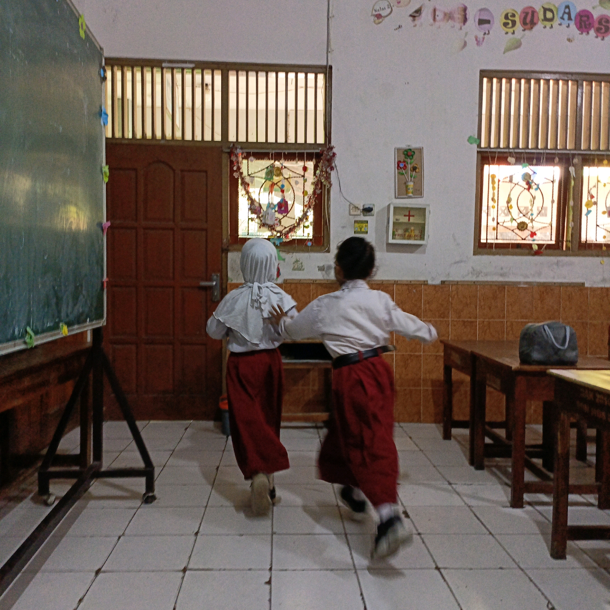 The image depicts the interior of a classroom in Salatiga, Indonesia. Two children, a girl wearing a white hijab and a red skirt, and another child also in a white top and red skirt, are shown from behind running towards the right of the frame. The child in the hijab is on the left and the other child is to her right. They appear to be moving in unison. The classroom features a dark green chalkboard on a stand to the left, a wooden door with a handle to the left of center, and two windows with wooden slatted shutters and decorative window hangings above the door and to the right. The walls are tiled with orange-brown squares up to window height, and white above. A metal first aid box is mounted on the wall to the right of the window. Wooden tables and benches are visible in the foreground and midground. The floor is tiled with square white tiles. The lighting suggests it may be daytime, with light entering through the windows. Decorations, including a banner with colorful letters spelling "SUDARS" and other colorful shapes, are visible on the upper part of the wall.