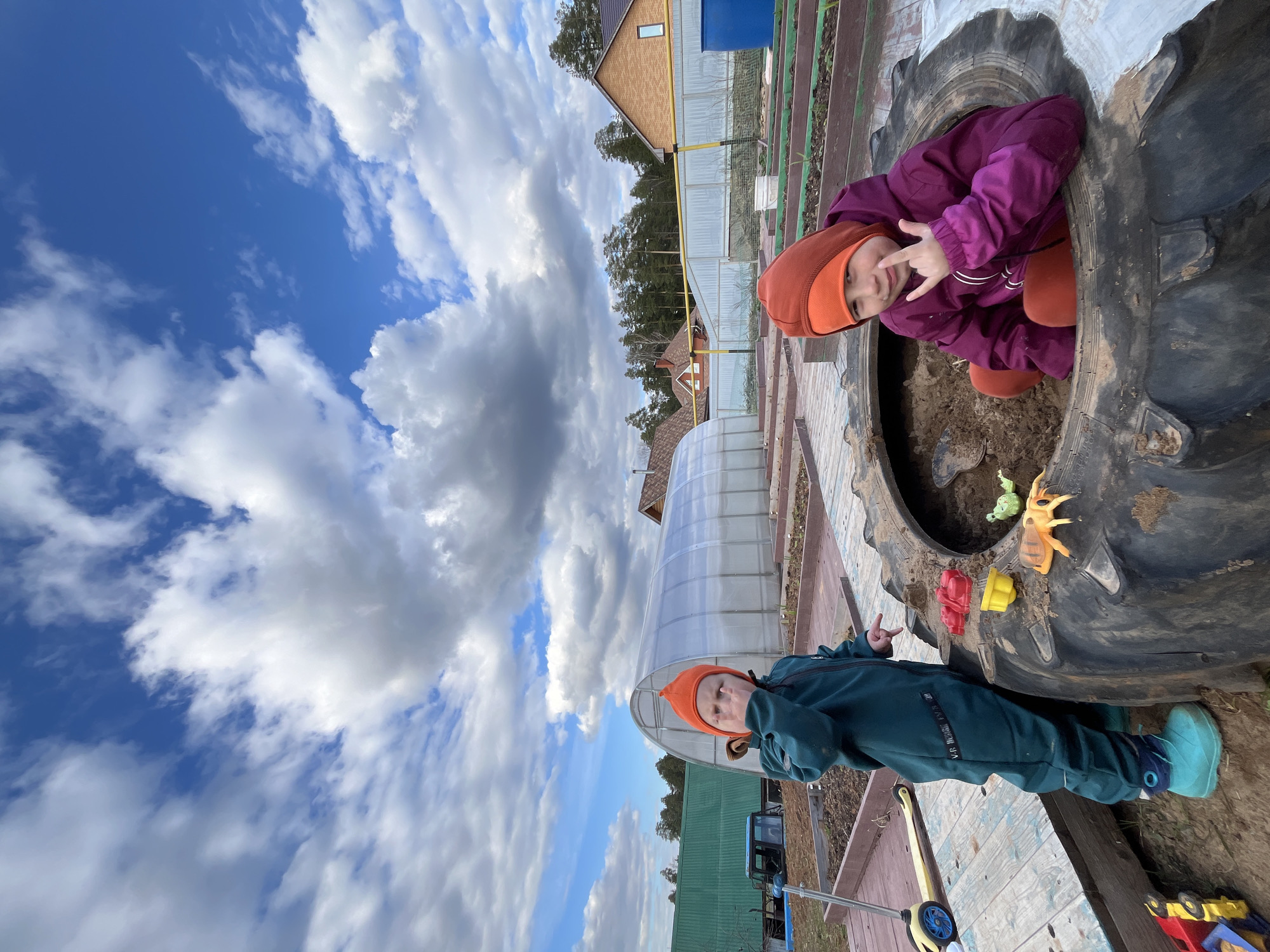The image, rotated 90 degrees clockwise from its likely natural orientation, depicts an outdoor play scene with two young children in Chaikovsky, Russia. The vast sky, a vibrant blue interspersed with large, fluffy white and grey clouds, dominates the upper-left portion of the frame.
In the lower-right, two children are engaged in play. One child, dressed in a purple jacket, orange pants, and an orange hat, is nestled inside a large, black tractor tire repurposed as a sandbox. This child looks directly at the camera, smiling and holding up a hand in what appears to be a "rock-on" or "peace" sign. Inside the tire, sand and a small black shovel are visible. Beside the tire, scattered toys include a green creature, an orange insect, and red and yellow plastic items.
Standing next to the tire on a wooden surface is the second child, wearing a dark green jumpsuit, bright turquoise boots, and an orange hat. This child also faces the camera, making a peace sign with one hand, while the other hand rests near their face. Unclear white text, possibly "VR_DESIGN", is visible on their jumpsuit. A yellow toy with wheels lies on the wooden surface nearby.
The background, visible along the vertical right side of the rotated image, reveals a typical countryside or dacha setting. Several houses with brown roofs and one with an orange-yellow facade are visible, along with a translucent arched greenhouse. A prominent yellow gas pipe runs horizontally across the scene. Beyond the structures, a line of trees or forest can be seen. The overall impression is one of joyful outdoor play on a bright, cloudy day.