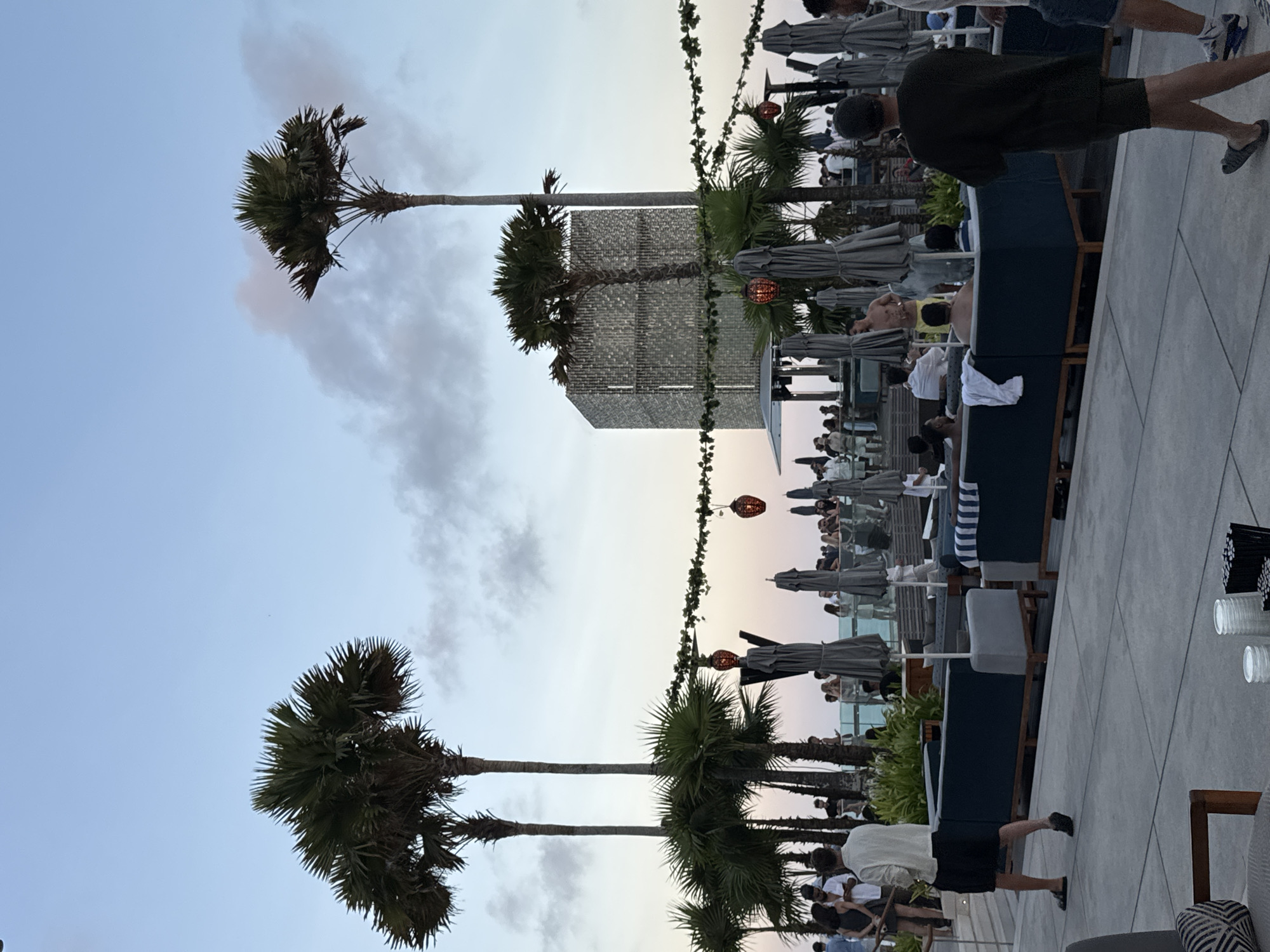 The image depicts a lively rooftop scene at dusk, likely a bar or restaurant, with people gathered and enjoying the evening. Tall palm trees frame the view, their fronds silhouetted against a pale sky. A modern, cube-shaped structure, possibly a building or architectural feature, stands in the mid-ground, its surface textured with what appears to be small tiles or panels. String lights with decorative red lanterns are strung across the scene, adding to the ambiance.
People are seated at tables and lounging on outdoor furniture, some under umbrellas. The atmosphere seems relaxed and social, with individuals dressed in casual attire. The time of day is indicated by the soft, fading light of sunset, creating a warm glow. The weather appears to be clear, with a few wispy clouds present. The combination of palm trees, open-air seating, and the twilight sky suggests a tropical or coastal location, consistent with Kuta Selatan, Indonesia. There is no visible text in the image.
