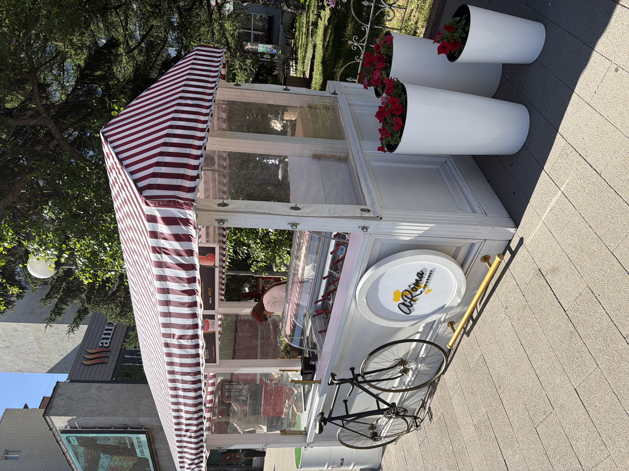 The image captures a street-side gelato cart adorned with a red and white striped awning, set against a backdrop of urban buildings and trees. The cart itself is white and features a transparent enclosure revealing tubs for gelato. A person in a red headband and white shirt is visible behind the counter. To the left of the cart, a black bicycle with two wheels is parked, its rear wheel close to the cart's side. The cart has a circular logo that reads "A'Roma GOURMET." In front of the cart, on the right side, are three tall white planters containing red flowers. The ground is paved with light-colored rectangular tiles. In the background, a building with a large digital billboard is visible, as is a prominent sign with the Cyrillic letters "АШАН". This street scene is located in Krasnoyarsk, Russia, likely near an "Auchan" supermarket given the signage. The lighting suggests it is daytime, with shadows cast by the cart and surrounding objects.