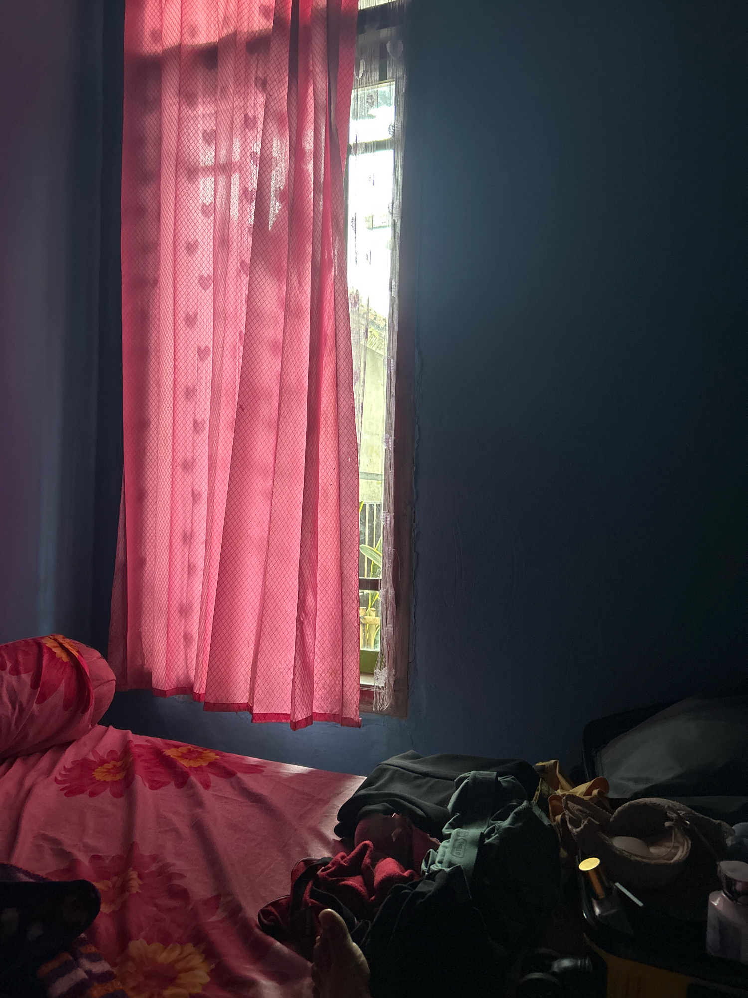 The image depicts a dimly lit bedroom interior in Cihaurbeuti, Indonesia. The scene is dominated by a blue wall and a pink curtain with a subtle heart pattern, which is partially drawn across a window. Sunlight streams through the window, illuminating the curtain and casting shadows. In the foreground, a bed with a pink floral patterned sheet is visible, with a pillow at the head. Several items of clothing, including dark-colored garments and a red cloth, are scattered on the bed and on the floor near the bed. A suitcase is also present, with some items spilling out. A person's foot is visible on the right side of the bed, suggesting someone is resting or sleeping. The overall impression is one of a lived-in, somewhat cluttered personal space.