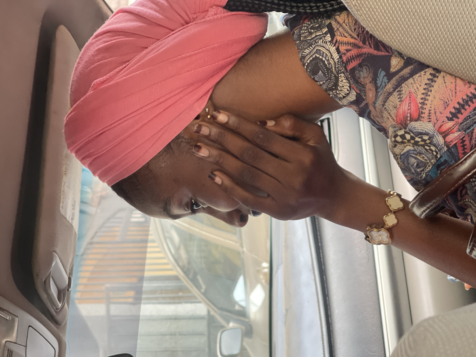 A medium shot captures a Black woman wearing a pink headscarf and a vibrantly patterned blouse, seated inside a vehicle. Her right hand rests on her chin, and she is looking out of the vehicle's window. Her nails are painted with a French manicure. A bracelet with clover-shaped charms adorns her left wrist. The interior of the car is visible, including the car's ceiling with a light fixture and a handle. Outside the window, another vehicle and a building with vertical slats are discernible. The scene appears to be set in Abuja, Nigeria, given the context provided.