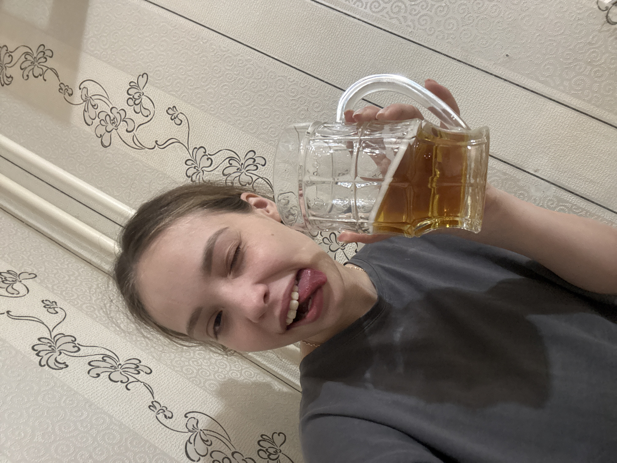 A young woman is holding a clear glass beer stein filled with a golden-colored liquid, presumably beer. She is sticking out her tongue and winking. The background features a patterned wallpaper. This image was taken indoors in Chaikovsky, Russia.