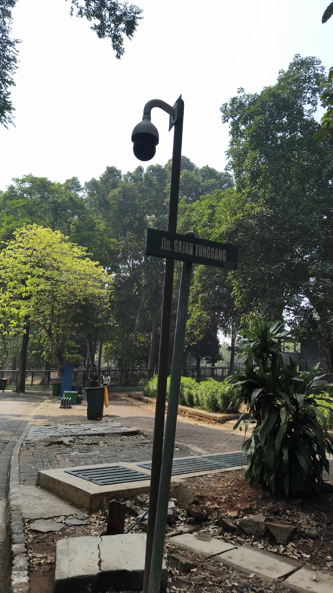 The image depicts a street sign that reads "Jln. Gajah Tunggang" in a park-like setting, possibly in South Jakarta, Indonesia. The sign is mounted on a pole that also supports a security camera. In the background, lush green trees and foliage are visible, indicating a natural environment within an urban area. Several people are present in the background, engaged in casual activities. The foreground features a paved walkway with some broken concrete slabs and debris, suggesting ongoing maintenance or wear and tear. The overall scene captures a common public space with elements of security, greenery, and human presence.