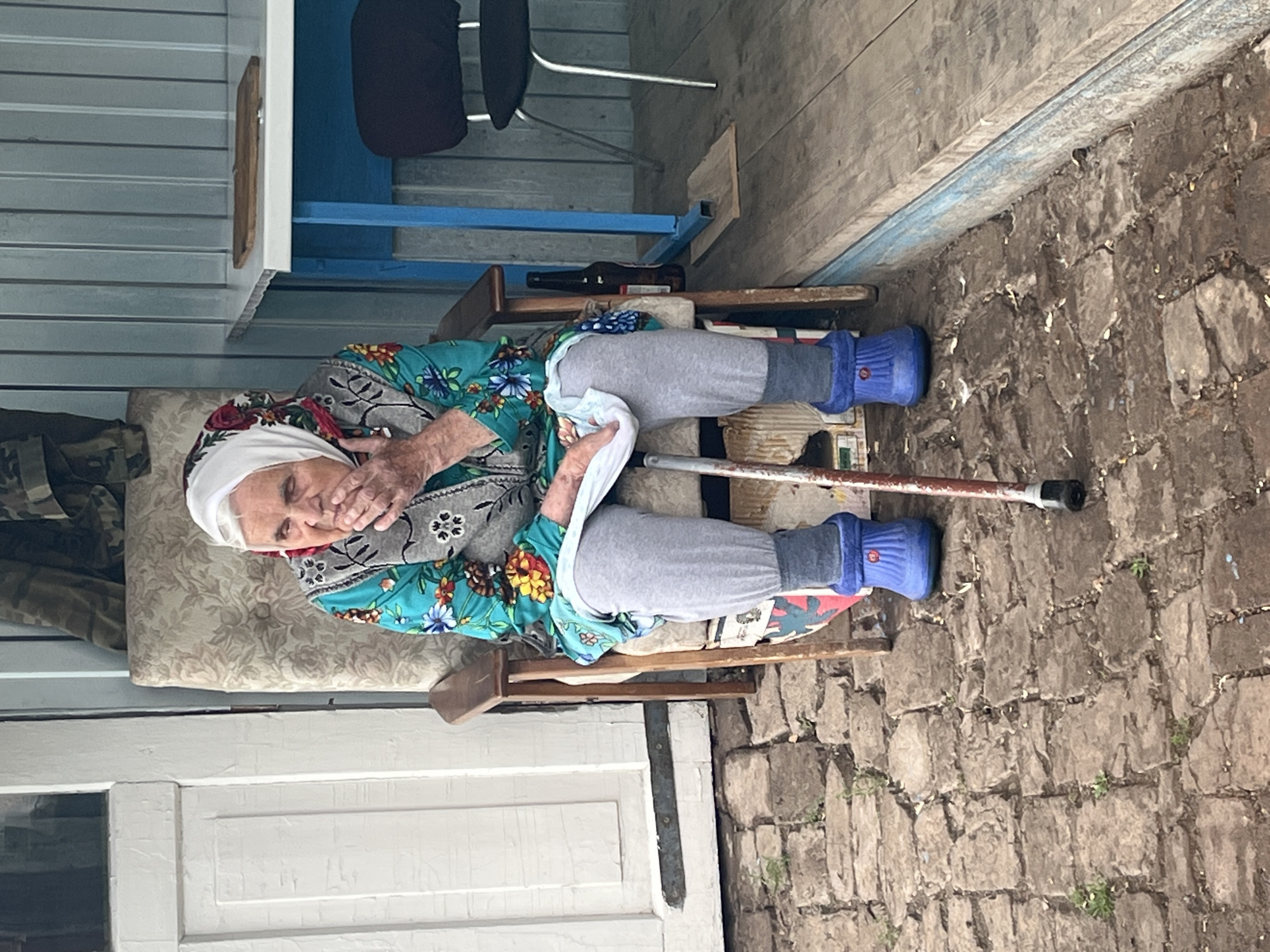 An elderly woman, wearing a white headscarf and a floral-patterned blouse with a grey embroidered vest, sits on a chair. She has her hand covering her mouth and appears to be either coughing or speaking. The woman is wearing grey pants and blue boots. A cane with a black handle rests beside her, its tip on the stone ground. The setting appears to be outdoors, possibly a porch or patio, with a wooden floor and a brick or stone wall in the background. To the left, there's a white door and part of a building structure with corrugated siding. A blue shelf unit with a dark chair is visible behind the woman. A brown bottle lies on a shelf within the blue structure. The overall scene suggests a casual, perhaps domestic, outdoor setting. The lighting and shadows imply it is daytime.