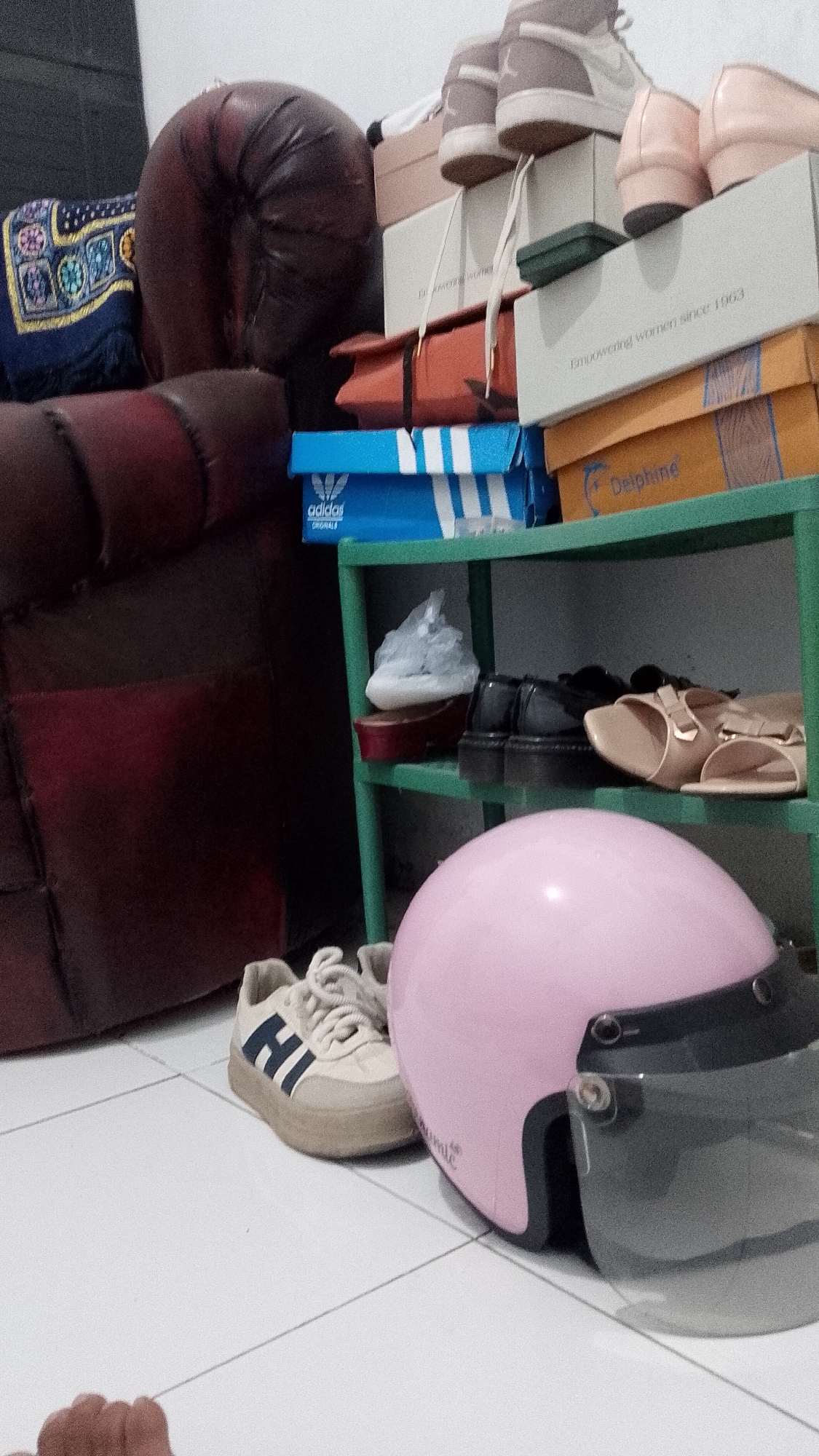 A low-angle, medium shot captures a cluttered indoor scene, likely a room in Karangkedawung, Indonesia, with a focus on footwear and storage. In the foreground, a pink helmet with a clear visor rests on a white tiled floor, next to a beige sneaker with blue stripes and white laces. To the left, a portion of a dark red sofa is visible. Behind the sofa, a stack of boxes and shelves are laden with shoes and other items. The shelves, made of green metal, hold several pairs of shoes, including high-heeled sandals, black platform shoes, and a white shoe wrapped in plastic. Boxes of various sizes and colors are stacked on top of each other, some with visible branding like "Adidas Originals" and "Delphine." The overall impression is one of domestic disarray, with an emphasis on shoe organization and storage.