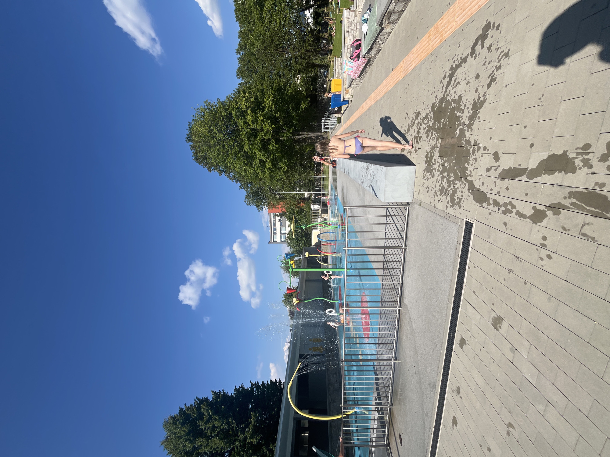 A young girl in a purple bikini stands on a concrete ledge, her body angled away from the camera, with a shadow cast on the wet ground beside her. She appears to be at a water park or splash pad on a sunny day. The water park has a shallow pool with colorful water features spraying water. A metal fence surrounds the pool area. In the background, trees, buildings, and other people can be seen. The sky is a clear blue with a few white clouds. The overall mood is one of leisure and fun in the sun. The image is taken from a high angle, looking down at the scene. There is no visible text in the image.