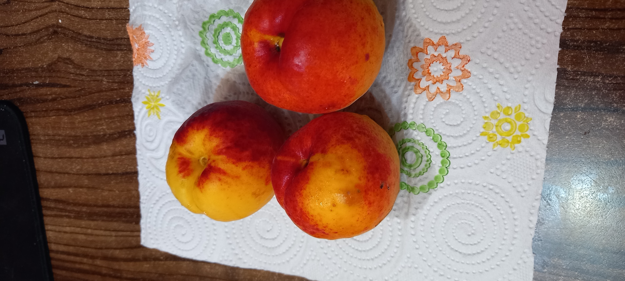 Three nectarines are arranged on a white paper napkin with floral designs. The nectarines are primarily yellow with significant red blush. The napkin is on a wooden surface. There is no indication of the city of Iskra, Bulgaria.