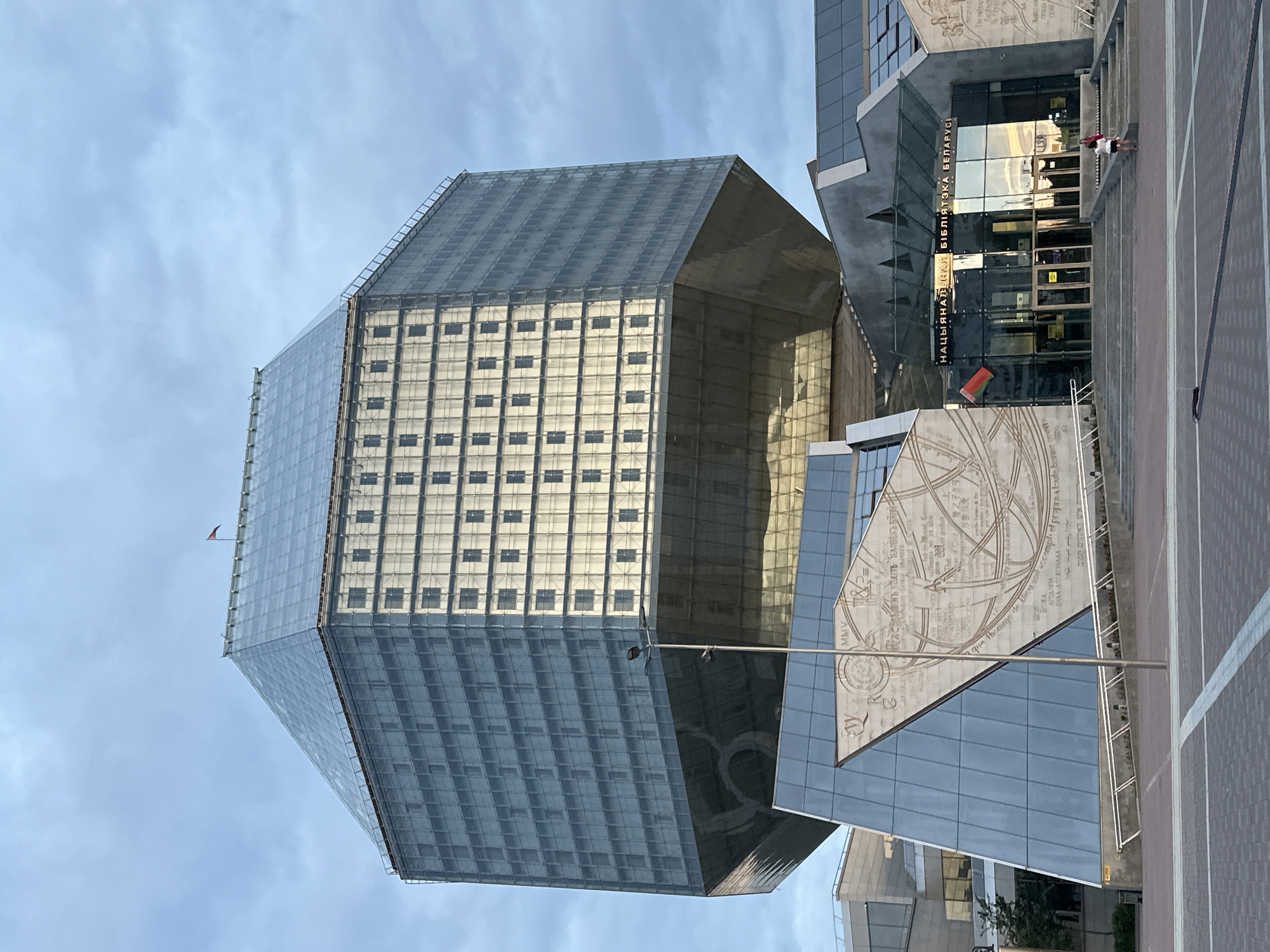 This image depicts the National Library of Belarus in Minsk, Belarus. The dominant structure is a large, multi-faceted building, architecturally rendered as a rhombicuboctahedron, clad primarily in reflective glass panels. The upper sections exhibit a bluish-grey hue from the glass, with one prominent side displaying a grid of numerous small, recessed square windows in a lighter, more reflective paneling, while another side features larger, darker glass panels. A small, dark red flag is affixed to the apex of the building's multi-faceted, sloping roof, which also consists of reflective material. The main structure rests upon a broad, angular, reflective base that creates a significant overhang.
Below the main structure, the entrance area consists of several angular, modern architectural elements featuring extensive glass and grey-toned cladding. Above the central glass entrance doors, the text "НАЦЫЯНАЛЬНАЯ БІБЛІЯТЭКА БЕЛАРУСІ" (National Library of Belarus) is displayed in Cyrillic script. A small Belarusian flag, featuring red and green with a traditional ornamental pattern, is visible to the left of the entrance. Flanking the entrance are large, light-colored relief carvings or murals on angled wall sections. These intricate designs include diagrams, what appears to be a map or celestial chart, and text in multiple languages, including Cyrillic, Latin (with a discernible phrase like "Que l'homme de Dieu soit accompli et propre à toute bonn..." and the Roman numeral "MMV"), Arabic, and East Asian scripts.
A wide set of shallow steps leads up to the main entrance. Two individuals are present on these steps to the right of the entrance: an adult, likely female, in a white top and dark shorts, and a child in a dark top and light shorts, both standing near the bottom of the steps and facing the building.
The foreground consists of a large, flat, paved plaza with a grid pattern formed by varying shades of grey paving stones and a single white dividing line. A grey lamppost stands vertically in the mid-ground, slightly to the left of center, obscuring a small part of the building's base. The sky is overcast and pale blue-grey, indicating diffused natural daylight. Minimal greenery is visible on the far left.