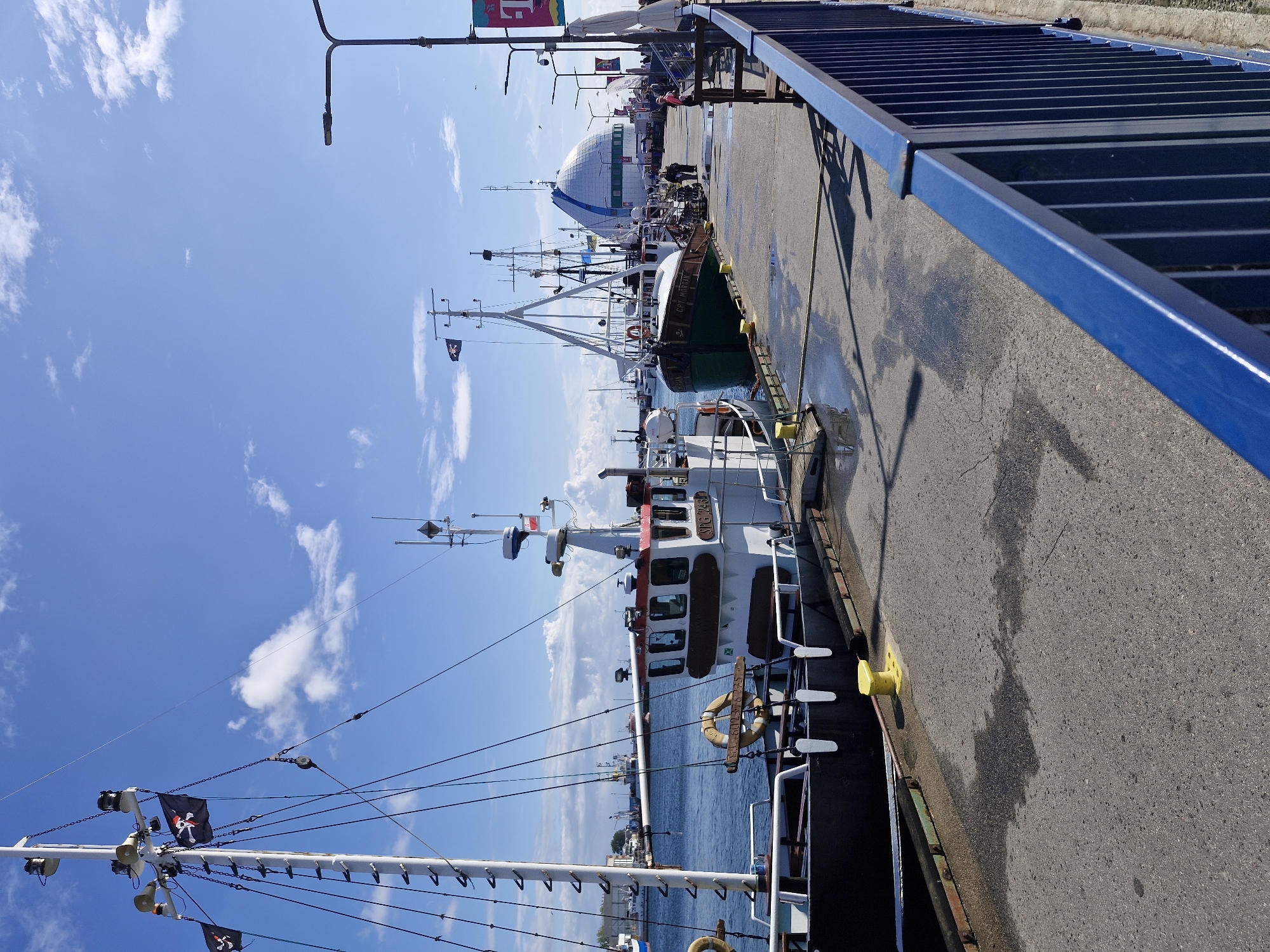 The image shows a bright, sunny day at a harbor in Hel, Poland, with several boats docked along a pier. In the foreground, a black and white fishing boat is moored, with a red stripe around its upper structure. Two life preservers are visible on its deck. Further down the pier, a green and white boat named "CAPTAIN MORGAN" is docked. In the background, a large, domed building with a blue and white striped exterior stands against a clear blue sky with scattered white clouds. Several other boats are visible in the harbor, and people are walking along the pier. A bright blue metal railing lines the right side of the pier.