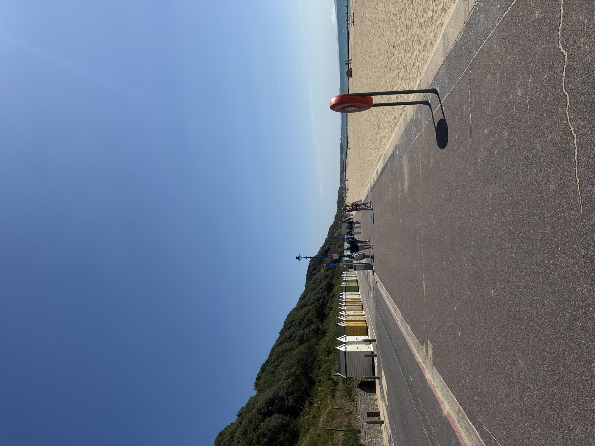 The image captures a sunny day on the promenade and beach in Bournemouth, United Kingdom. A paved walkway runs along the left side of the image, bordered by a road and a row of colorful beach huts. The beach huts are varied in color, with white, yellow, and green being prominent. Behind the beach huts, a green, treed hillside rises steeply.
On the promenade, several people are walking, appearing relaxed and enjoying the weather. They are dressed in casual attire, suggesting a typical day at the seaside. To the right of the promenade is a wide expanse of golden sand, the beach. In the foreground of the beach, a red and white life buoy on a stand is prominently positioned. Further down the beach, other beachgoers can be seen, some appearing to be sitting or standing. The sea is visible in the distance, a calm blue under a clear, bright sky. The overall scene conveys a peaceful and pleasant atmosphere of a popular seaside resort.