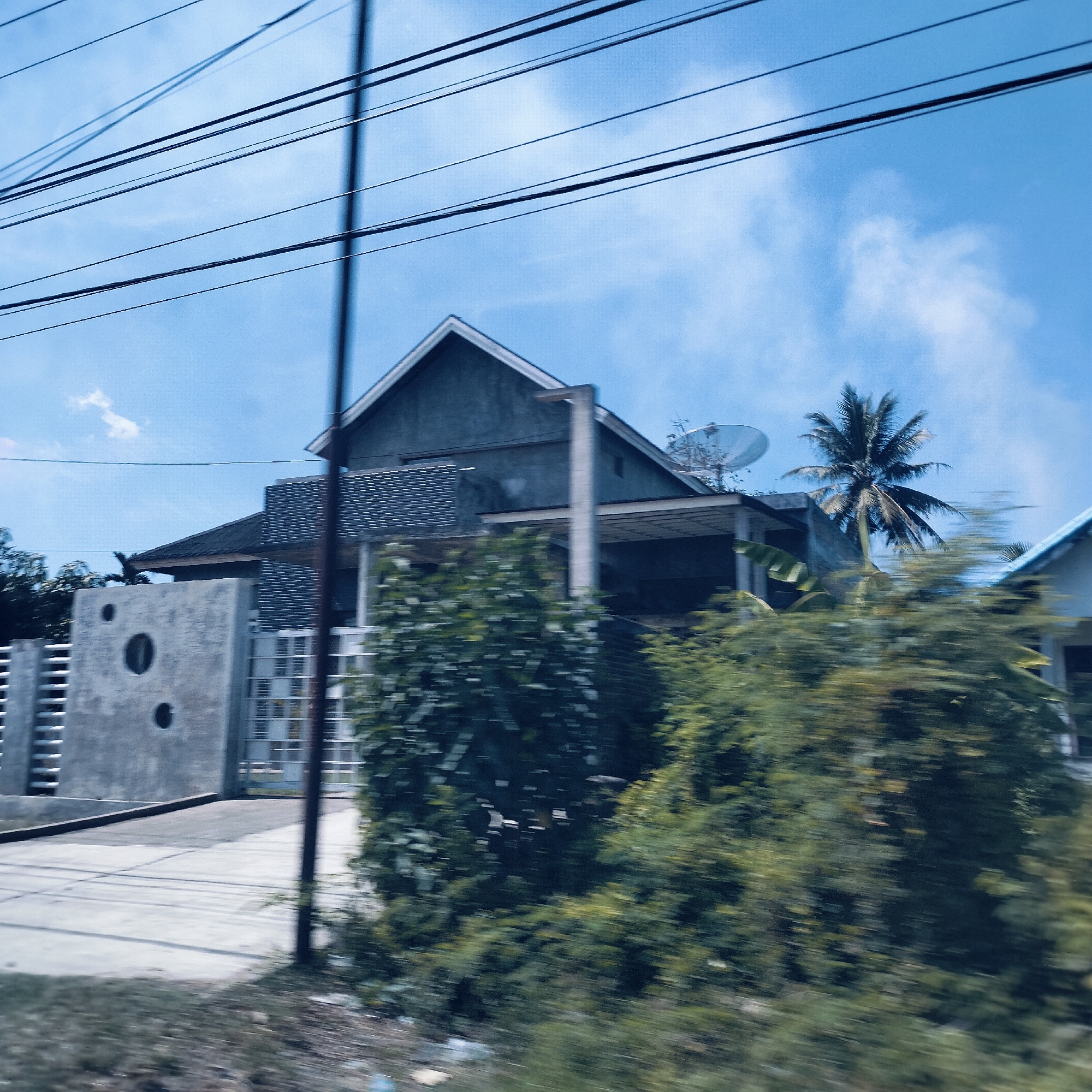 The image captures a residential scene in Keudelapang, Indonesia, taken from a moving vehicle, evidenced by the motion blur in the foreground. The prominent subject is a modern-style house with a grey, angular roof and concrete walls, situated under a clear blue sky with sparse white clouds. Multiple dark utility wires stretch across the upper portion of the frame.
The house features a covered patio or carport area and a satellite dish mounted on its roof. Lush green trees and bushes frame the house, with a tall palm tree distinctly visible behind it, suggesting a tropical environment. To the left of the house, a grey concrete fence or wall with distinct circular cutouts and a gate provides a boundary. A concrete driveway or access road leads towards the house from the foreground. A dark utility pole is partially visible on the right side of the driveway. The overall setting depicts a sunny daytime scene in a seemingly quiet, residential area. No specific activity or visible text is present.