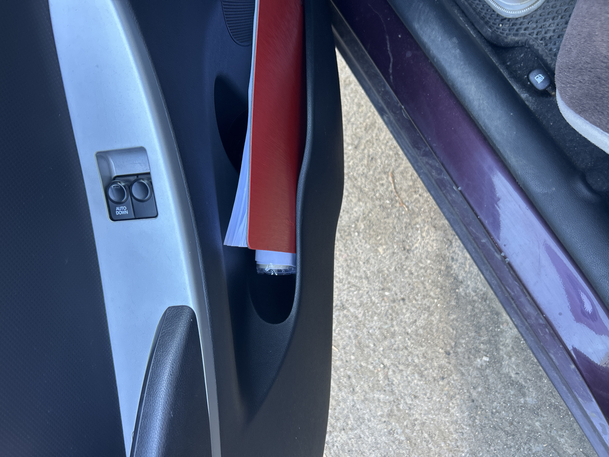 The image shows a close-up view of the interior of a car door, specifically the lower door pocket and the window controls. A red booklet or folder is tucked into the door pocket, partially obscuring some white papers. The window controls, labeled "AUTO DOWN", are visible on a silver trim panel above the pocket. The surrounding car interior is dark, and the exterior appears to be a paved surface. There is no information in the image that allows for a determination of the location in the city of Orenburg, Russia.