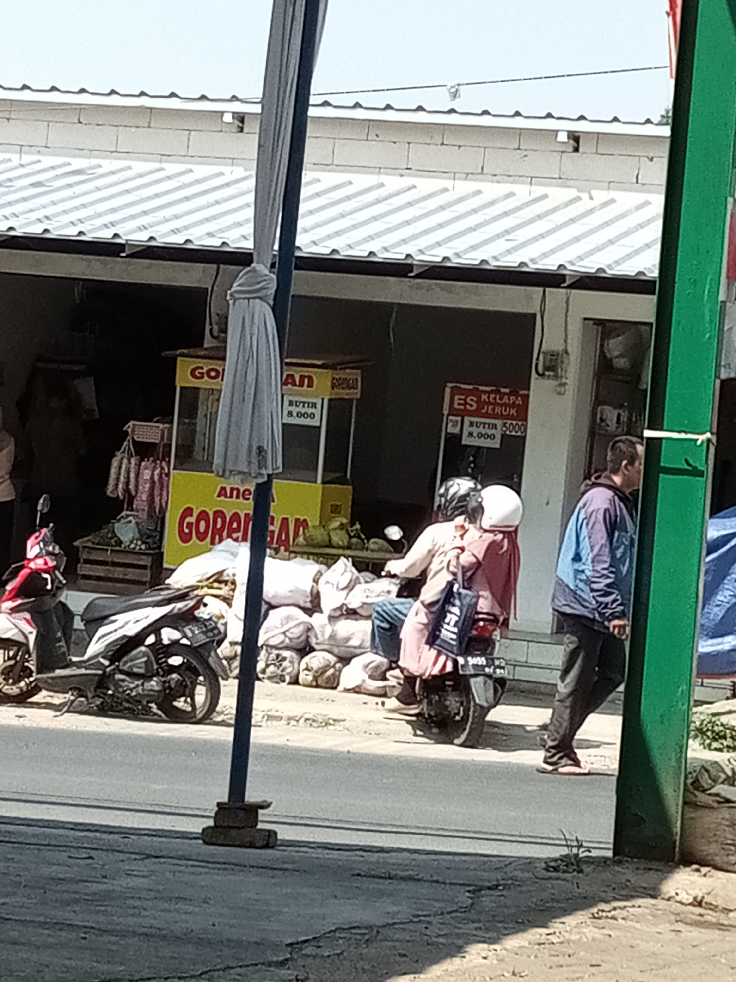 In Cipanjak, Indonesia, an outdoor scene unfolds with a focus on a roadside food stall. A yellow food cart with the signage "GORENGAN" (fried food) and "ANeka GORENGAN" (various fried food) is visible. Adjacent to the cart are stacks of what appear to be sacks, possibly containing goods. A white motorcycle is parked to the left, and another motorcycle with two people on it is positioned in front of the food cart. The person in the back is wearing a pink headscarf and a pink garment, while the person in the front is wearing a beige shirt and a helmet. A man in a blue and purple jacket and dark pants is walking away from the motorcycle. In the background, a white-tiled building with a corrugated metal roof stands. A green pole is prominently located on the right side of the frame. The ground is paved with asphalt, suggesting a street or road.