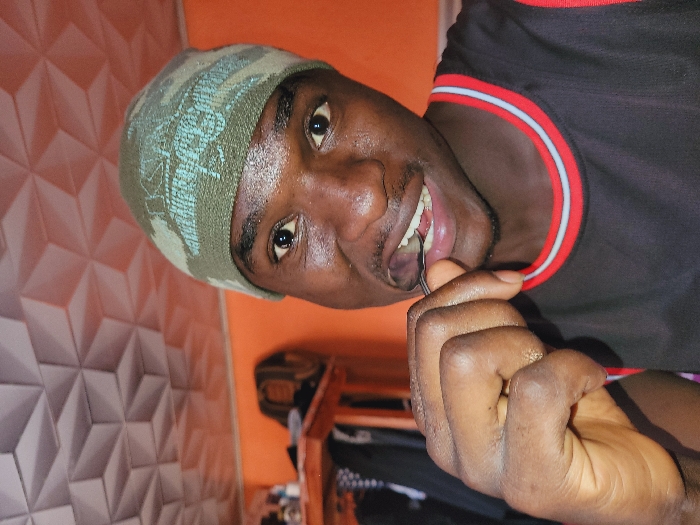 An adult male is depicted in an indoor setting in Bukuru, Nigeria. The individual, with dark skin, directly faces the camera. He wears a green and light blue camouflage-patterned beanie and a black sleeveless shirt with red and white collar trim. His right hand holds a small, curved metallic object, possibly a spoon or dental implement, which is positioned between his slightly open lips and teeth. A reflective sheen is visible on his forehead and cheeks. The background features a plain orange wall, topped by a ceiling with white or light gray three-dimensional geometric paneling. A partial view of a wooden surface and dark fabric is present on the left side of the frame.
