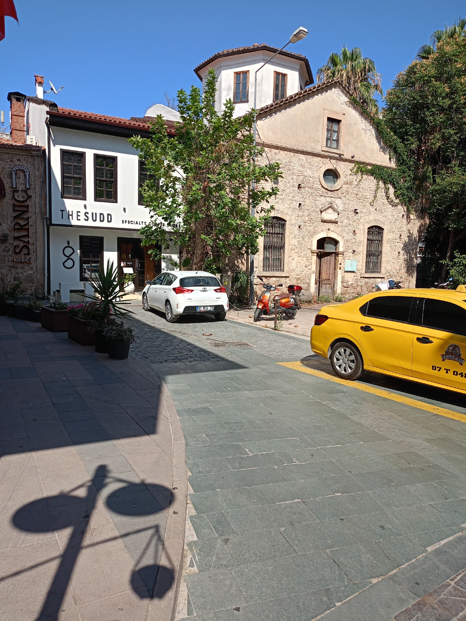 In Antalya, Turkey, on a sunny day, a yellow taxi with the license plate "07 T 048" is parked on a street paved with gray cobblestones, alongside a white car and an orange scooter. The taxi is in the foreground, and the street curves away from the viewer.
To the left of the street, a white building with dark windows and the sign "THESUDD SPECIALTY COFFEE STORE" stands. Next to it, a traditional stone building with arched windows and a weathered wooden door is visible. Greenery, including trees and potted plants, surrounds the buildings. The sky is clear and blue. A street light casts a long shadow across the cobblestones.