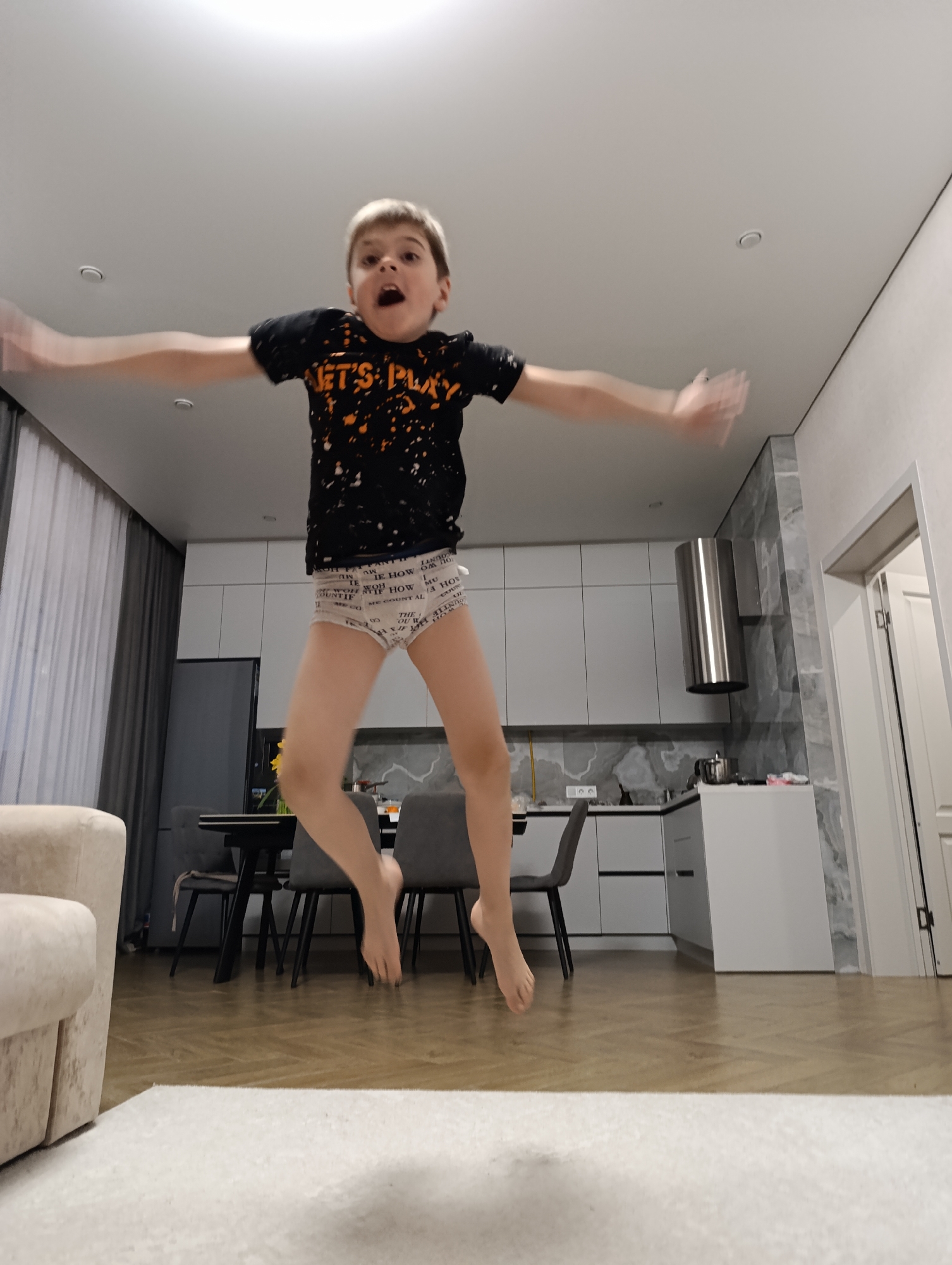 A young boy is jumping in the air with his arms outstretched in what appears to be the kitchen of a modern home. He is wearing a black t-shirt with an orange graphic that reads "LET'S PLAY" and patterned underwear. The kitchen features white cabinets, a stainless steel range hood, and a dining area with a black table and gray chairs. A tan sofa is visible on the left side of the frame, and a white rug is in the foreground. The location is presumably in the city of Chaikovsky, Russia, based on the prompt.