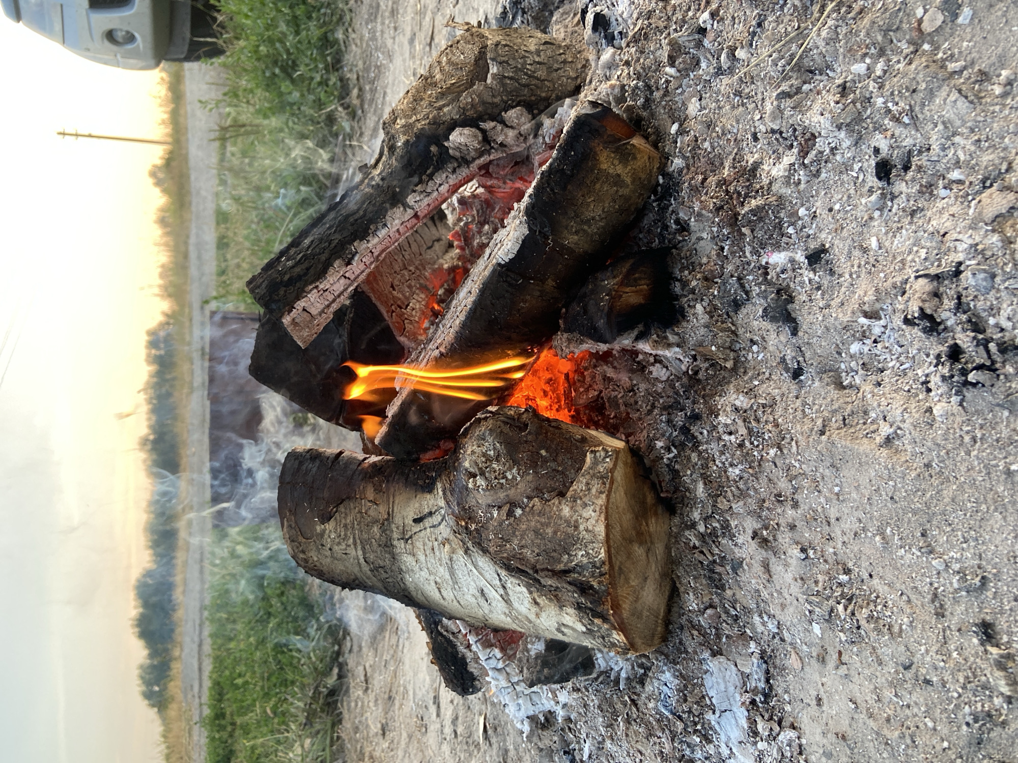 A campfire is burning outdoors on a sandy ground. The fire is made of several logs, some of which are partially burnt, and embers glow with orange and red light. Smoke rises from the fire. In the background, there is green grass and a dirt road. A vehicle is visible on the right side of the image, and the sky is a soft yellow and white, suggesting it is either sunrise or sunset. The location is identified as Peshcherka, Russia.