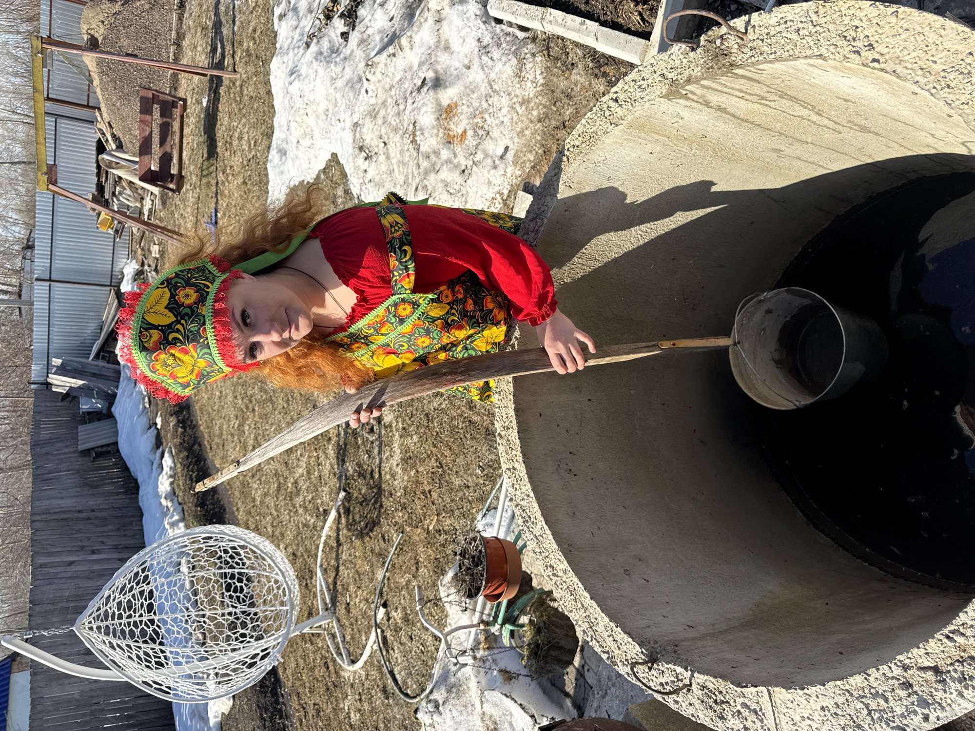 The image shows a woman dressed in traditional Russian clothing standing next to a concrete well. She is holding a wooden stick with a bucket attached to it. The well is circular and appears to be filled with water. The background includes a patch of melting snow, some wooden structures, and a white cage-like object. The location is in a rural or semi-rural area of Chaikovsky, Russia, as indicated by the traditional clothing and well design.