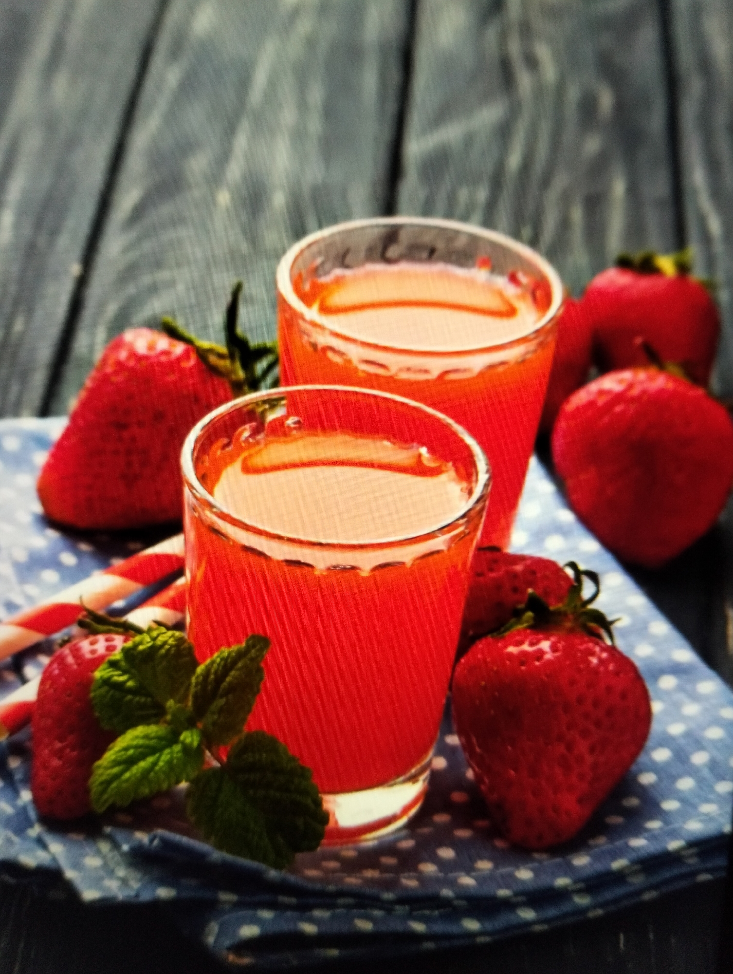 In the city of Payagaboh, Indonesia, on a rustic wooden surface, a refreshing scene unfolds. Two clear shot glasses, brimming with vibrant strawberry juice, stand as the centerpiece. Surrounding them are plump, red strawberries, their seeds glistening, adding to the allure. A few sprigs of fresh mint leaves rest on a blue and white polka-dotted cloth, their green adding a touch of contrast to the red. Two striped straws, in shades of red and white, peek out from behind the glasses, hinting at the enjoyment to come. The overall ambiance is one of summer refreshment and delightful indulgence.