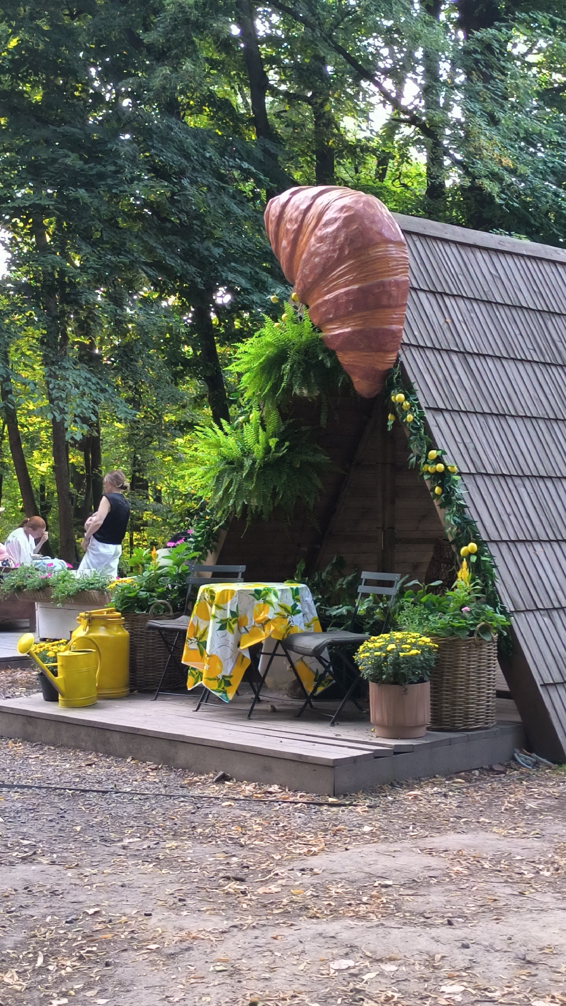 The image shows an outdoor seating area set up in a park-like environment, possibly at the Lviv National Academy of Arts in Lviv, Ukraine. A large, decorative croissant sculpture is prominently displayed on top of a triangular wooden structure that serves as a backdrop. In the foreground, a wooden deck has a small table covered with a yellow tablecloth featuring lemon prints. Two chairs are placed around the table. To the left of the table, two yellow watering cans and a large yellow canister are situated on the deck. Further left, two individuals are partially visible; one is seated and appears to be looking down, while the other stands. Lush green ferns hang from the wooden structure. The ground around the deck is covered with gravel and fallen leaves. The area is surrounded by trees, suggesting a natural, shaded setting. The overall scene evokes a whimsical, outdoor cafe or exhibition space.