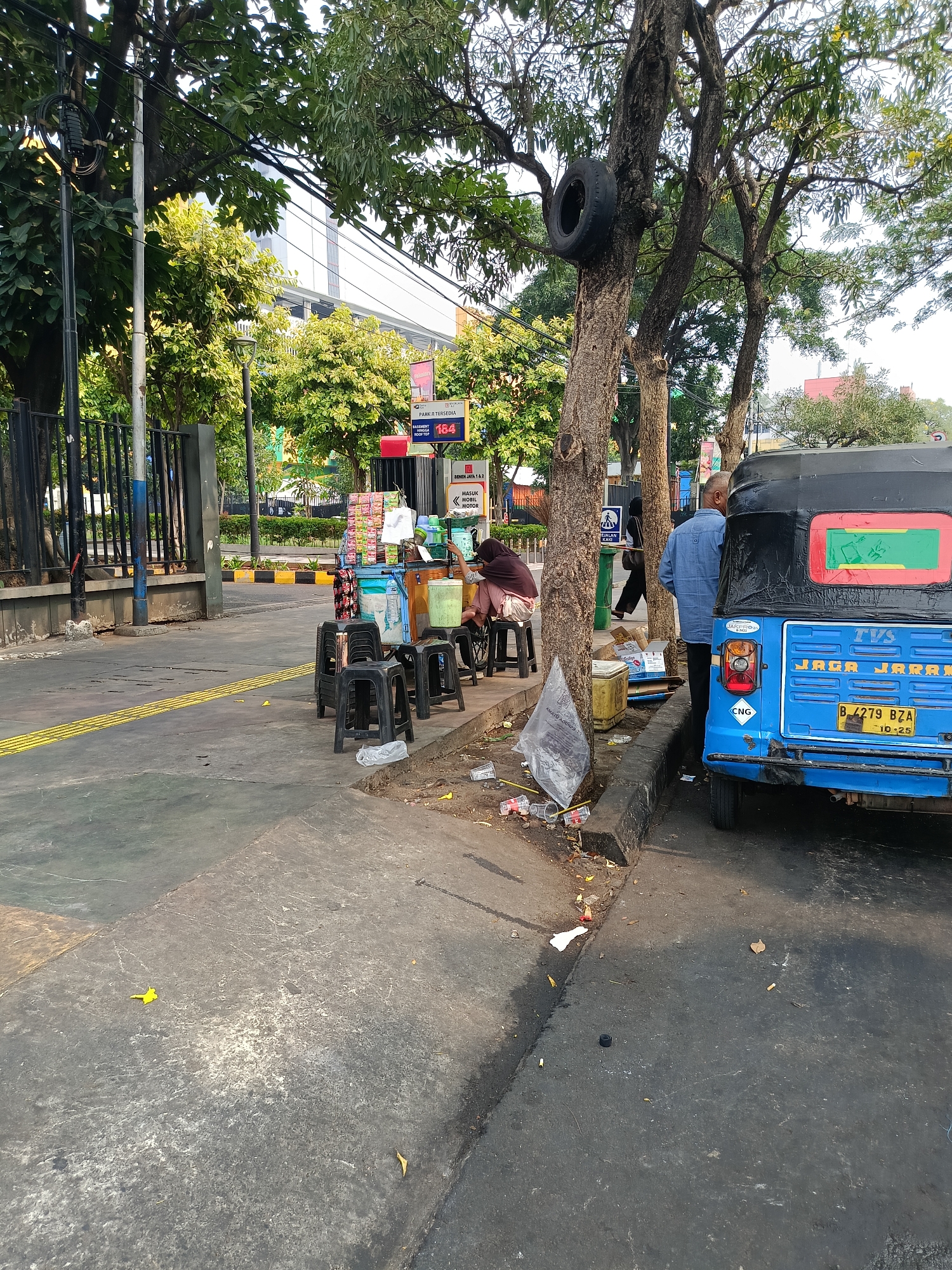 The image depicts a street scene in Central Jakarta, Indonesia, on a sunny day. In the foreground, a blue auto-rickshaw, identified by the license plate "B-4279 BZA", is parked on the right side of the frame. The auto-rickshaw has "TVS" branding and "JAGA JARAK" (keep distance) written on its back. Adjacent to the auto-rickshaw, there is a sidewalk lined with trees. One tree has a black tire mounted on a branch.
On the sidewalk, a small mobile cart displays various items, possibly for sale, with several plastic stools arranged around it. Two individuals are seated on these stools, seemingly interacting with the cart. A third person, wearing a blue shirt, stands near the auto-rickshaw, facing away from the camera. The sidewalk area around the cart and stools is cluttered with various items, including plastic containers, bags, and debris.
The street itself has distinct yellow tactile paving indicating a pedestrian pathway. In the background, a building with a glass facade and trees line the street. A black metal fence is visible on the left side, bordering a green area. Several signs are also present, including one with the number "184" and others with text in Indonesian. The overall impression is of a bustling urban environment with street vendors and daily life activities.